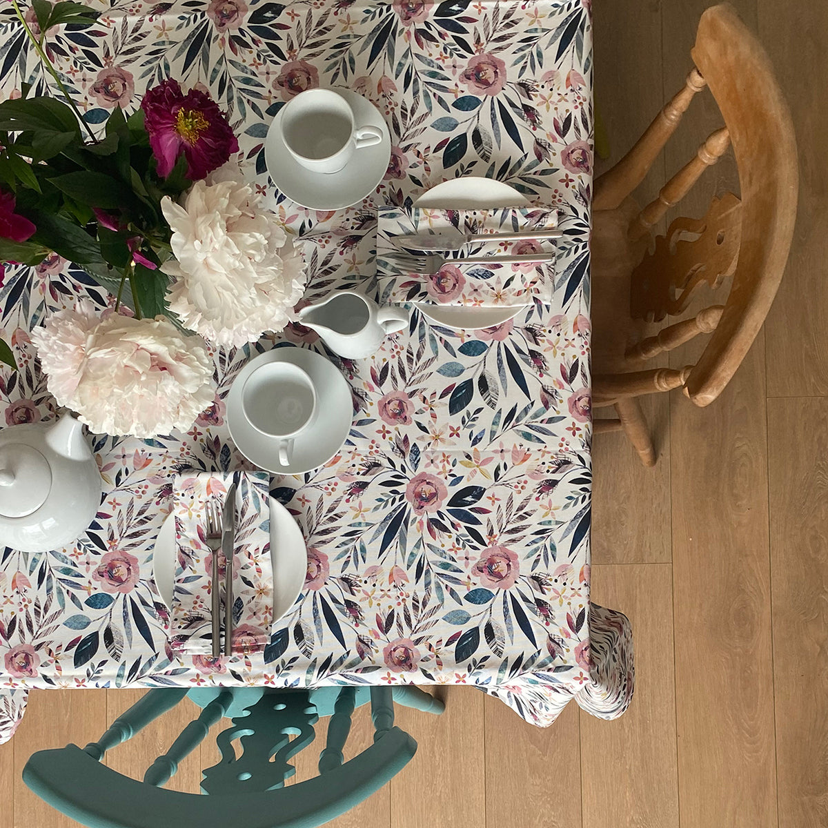 Summer Floral Table cloth featuring textured dusky pink roses and green foliage on a white background.  The table has been set with 2 chairs and two small side plates and a tea pot and cups. a vase of flowers in in the middle of the table.