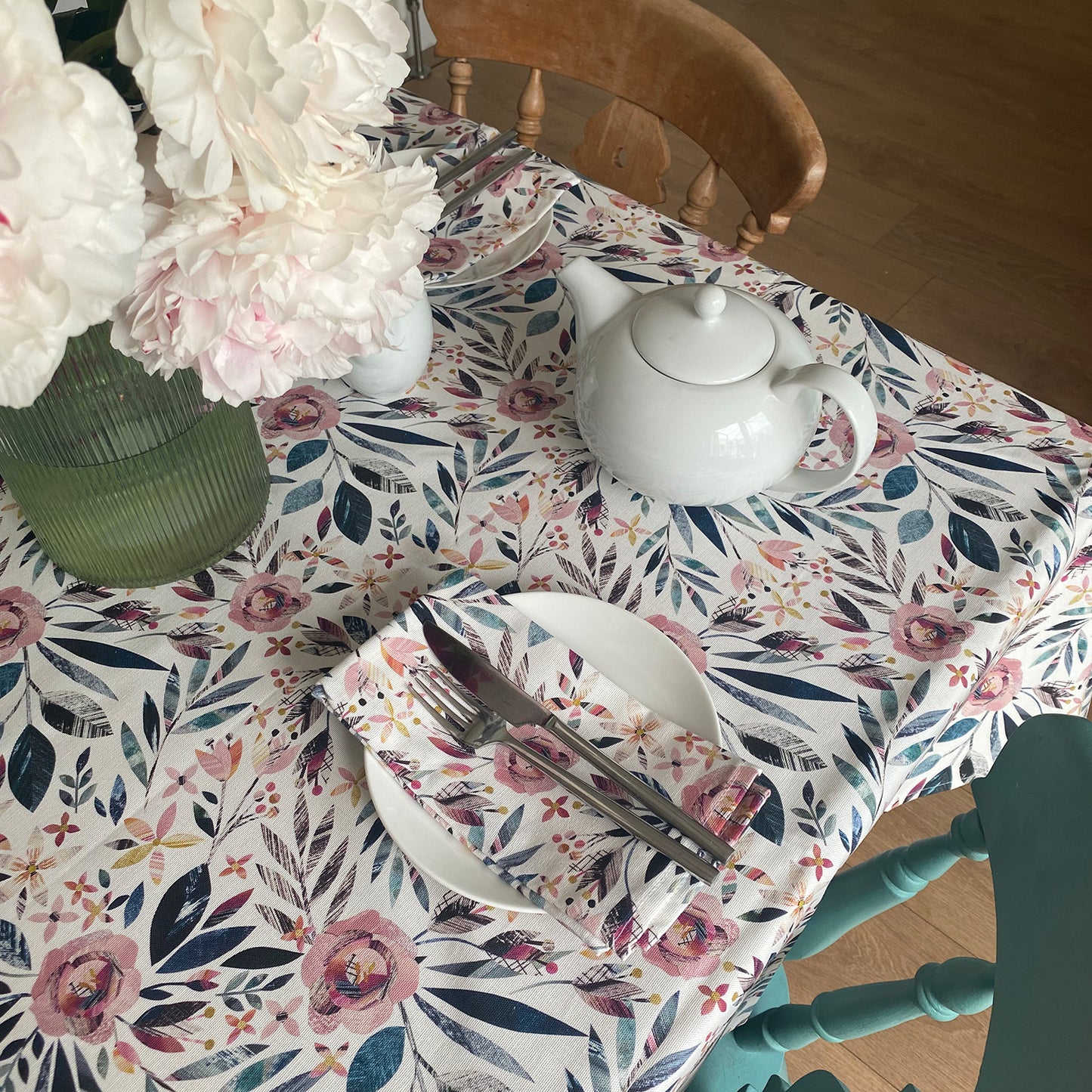 Summer Floral Table cloth featuring textured dusky pink roses and green foliage on a white background. The table has been set with 2 chairs and two small side plates and a tea pot and cups. a vase of flowers in in the middle of the table.