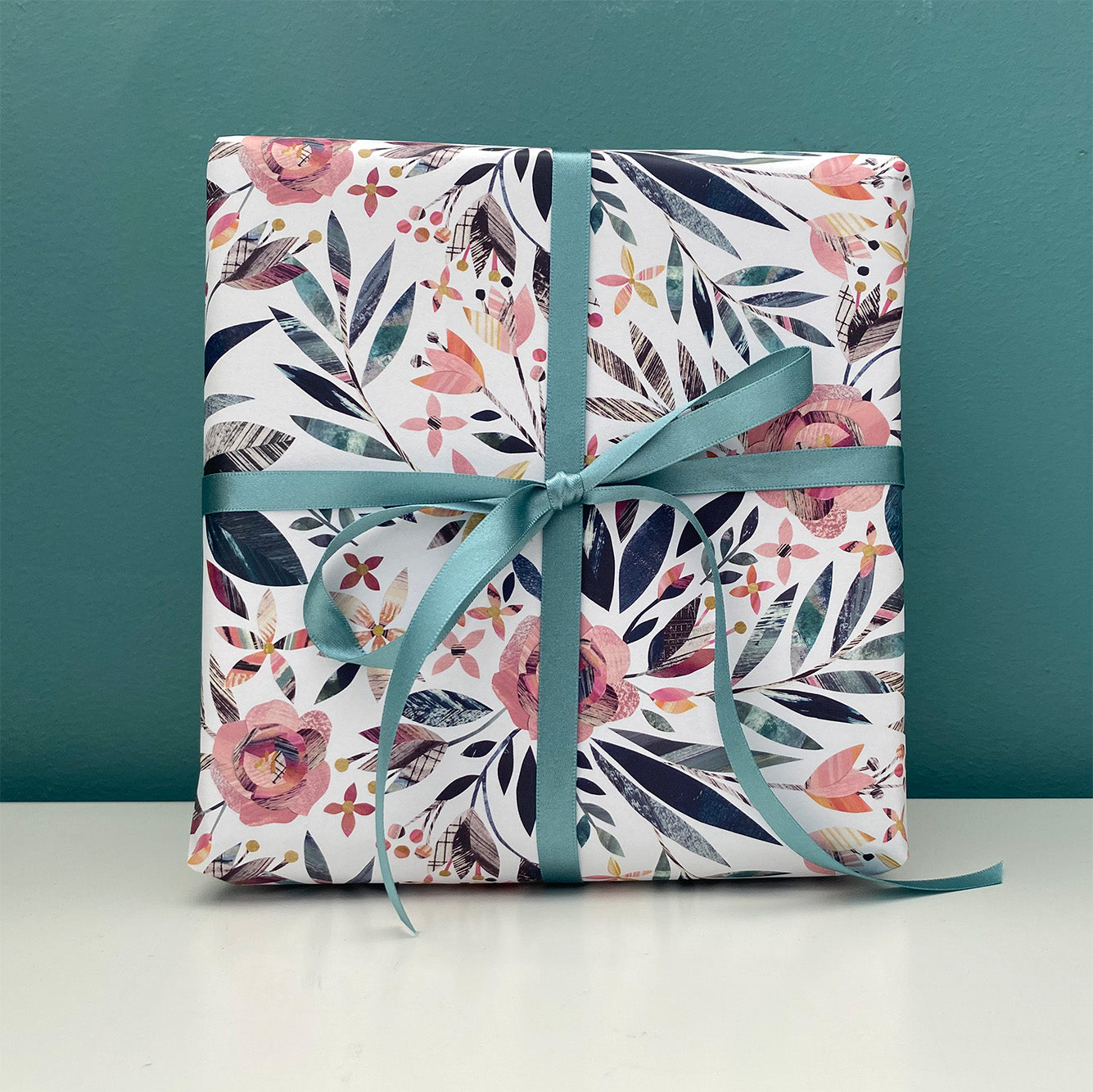 A present has been wrapped in the Summer Floral wrapping paper featuring dusky pink roses with green leaves. It has been tied with coordinating green silk ribbon in a bow.  The present has been placed on a white shelf against a blue green wall.