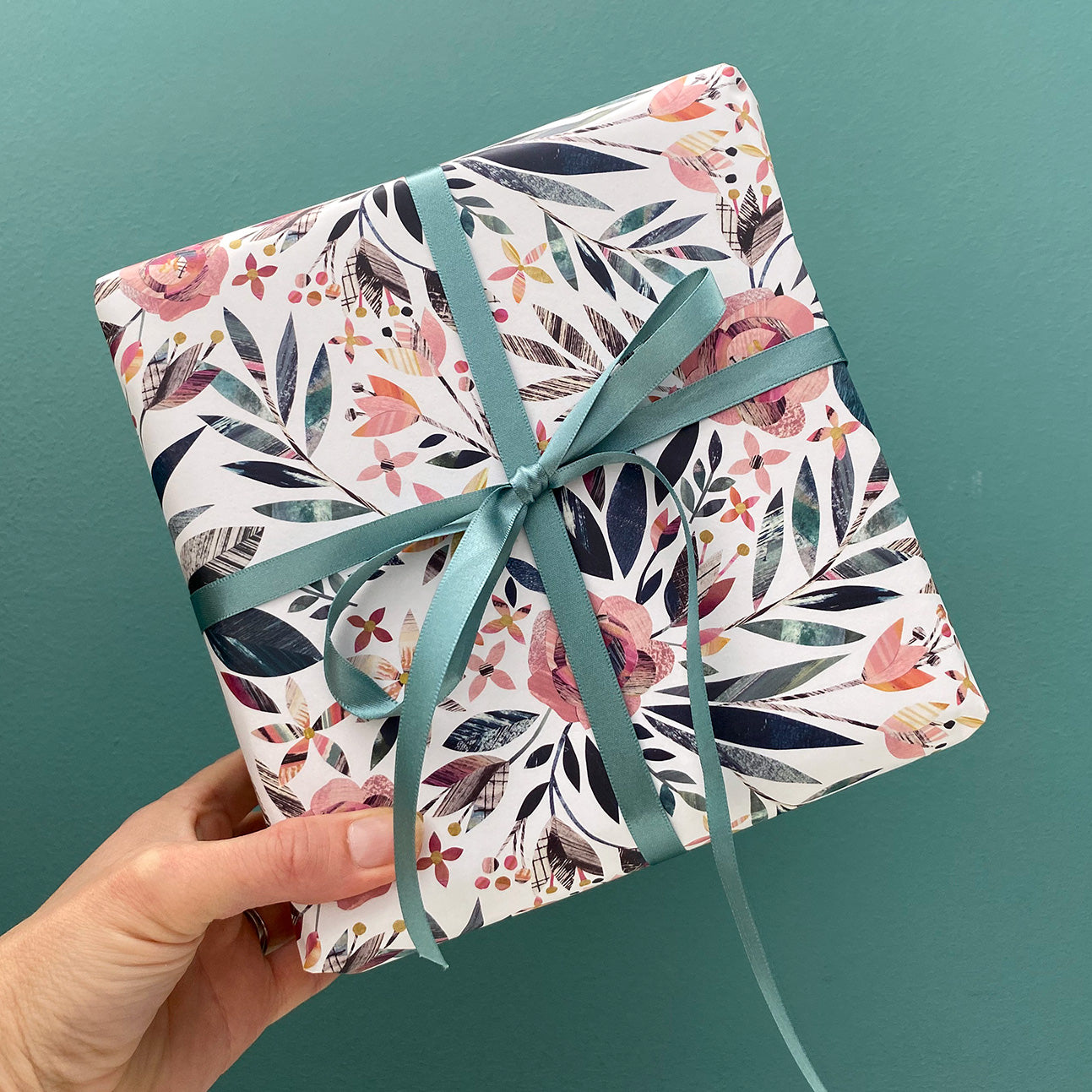 A present has been wrapped in the Summer Floral wrapping paper featuring dusky pink roses with green leaves. It has been tied with coordinating green silk ribbon in a bow.  The present is being held against a Blue green background.