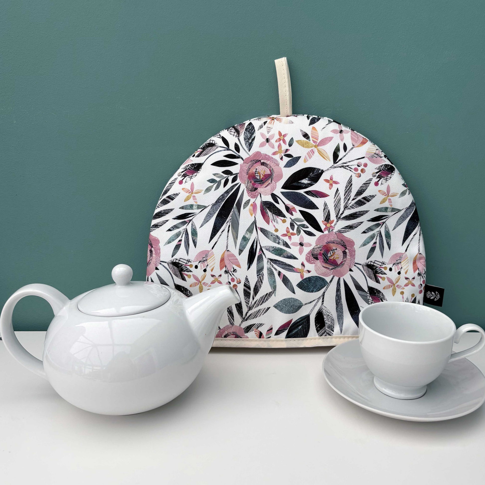 A Summer Floral Tea Cosy has been placed on a white shelf with a white tea pot, tea cup and saucer against a teal wall.