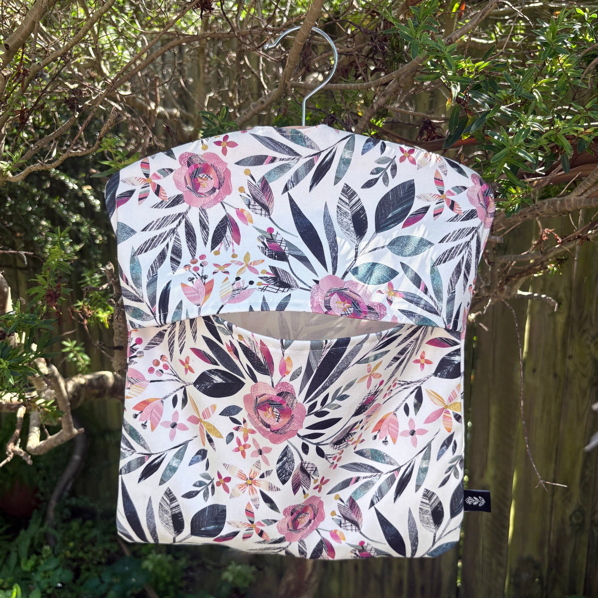 A Summer Floral patterned peg bag hanging outdoors on a wooden fence with greenery.