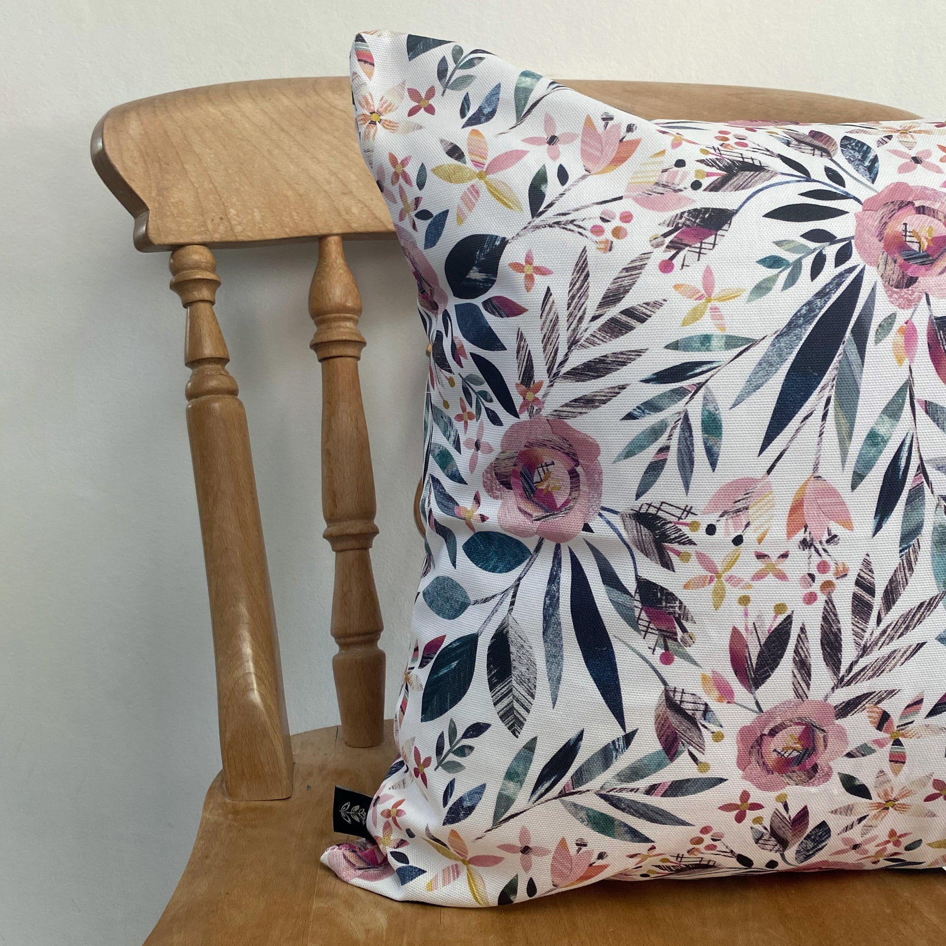 A Square Summer Floral Cushion, has been placed on a wooden chair against a pale grey wall.