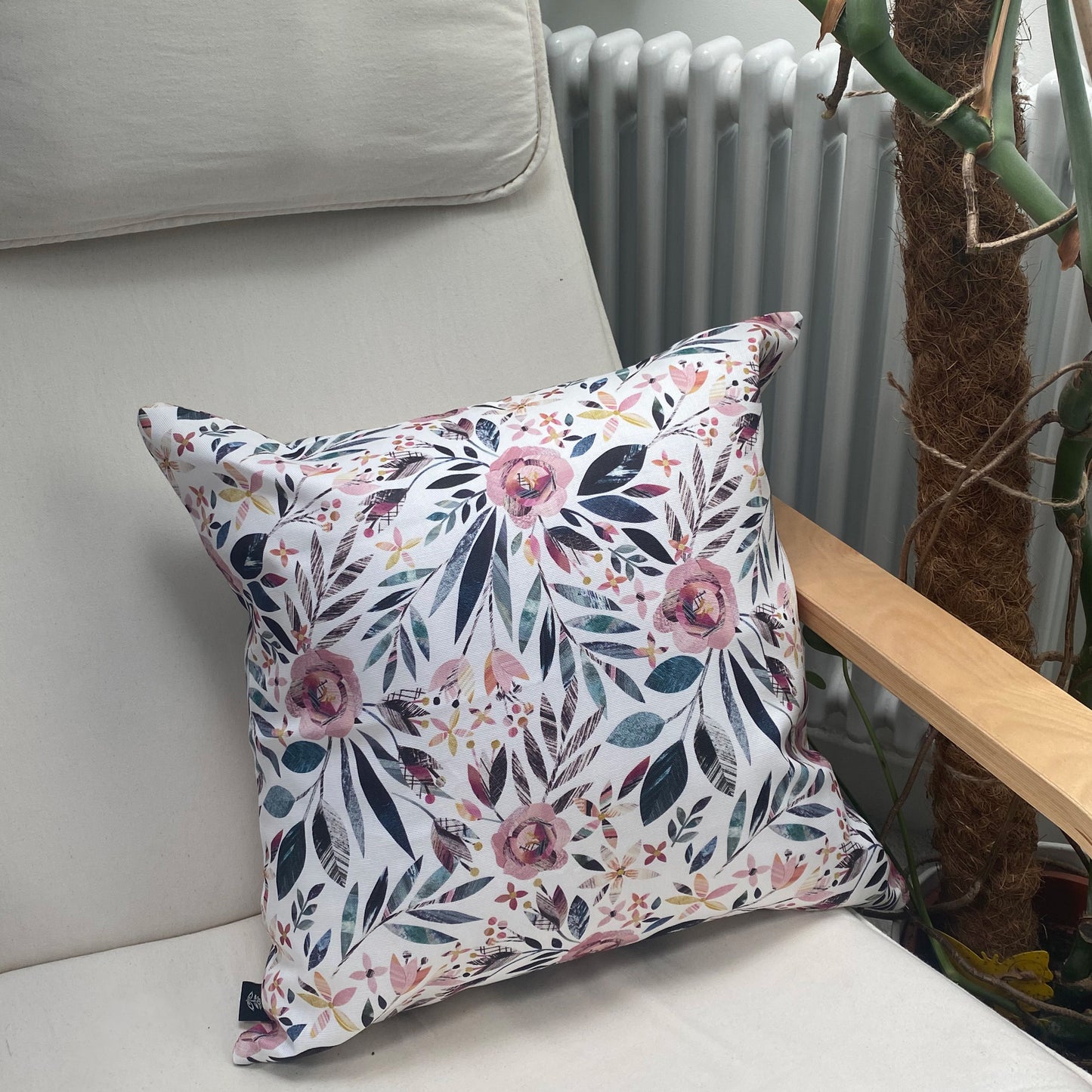 A square Summer Floral Cushion has been placed on a cream canvas and wooden arm chair.  A radiator and plant can be seen in the background.