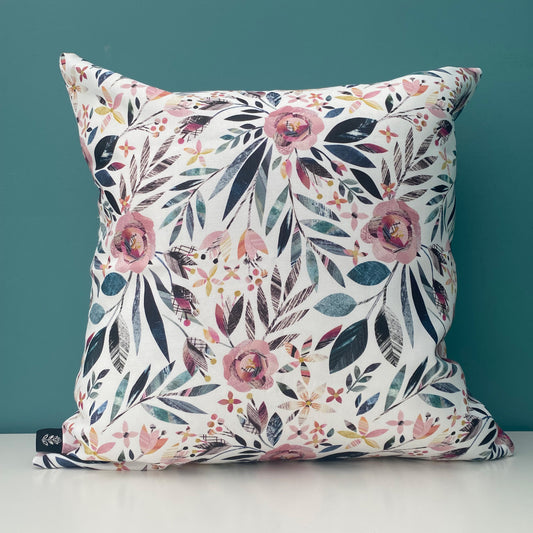 A square Summer Floral Cushion featuring textured dusky pink Roses and green leaves has been placed on a white shelf against a blue/green wall.