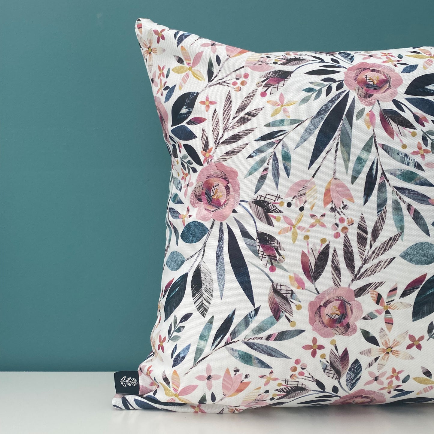 A close up of half of a square Summer Floral Cushion featuring textured dusky pink Roses and green leaves has been placed on a white shelf against a blue/green wall.