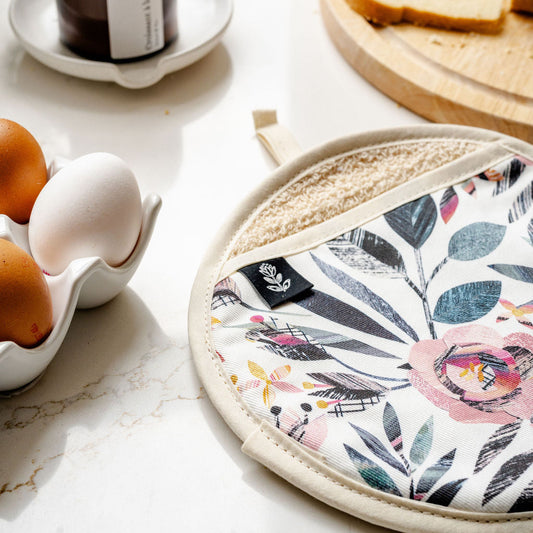 A round potholder with a floral and botanical print pattern in pink, green, and grey on a white background, featuring a beige cotton towelling lining and a cream fabric rim. It is styled on a white marble countertop alongside a candle, sliced bread, and eggs.