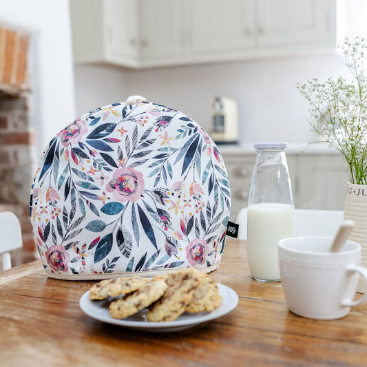A Summer Floral Tea Cosy has been placed on a wooden table in a kitchen with a plate of cookies, a up of tea and a small bottle of milk.