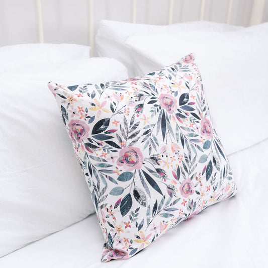 A square decorative throw pillow with a pink, green, and gray floral pattern, resting on a white bed with white pillows and a white metal headboard.