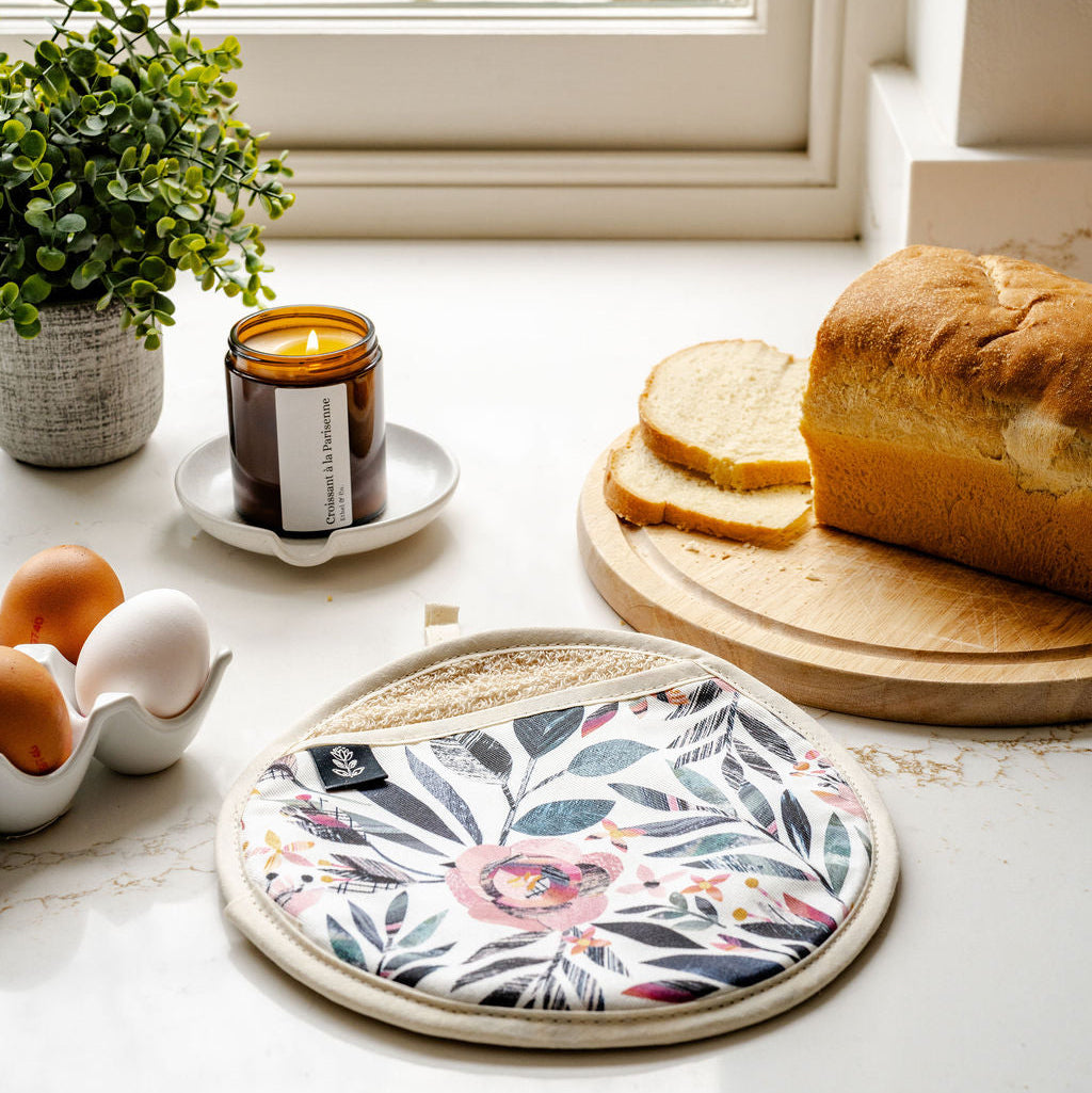 A round potholder with a botanical floral pattern in pink, green, and blue on a white background, styled on a kitchen counter next to a loaf of bread, eggs, a candle, and a small plant.