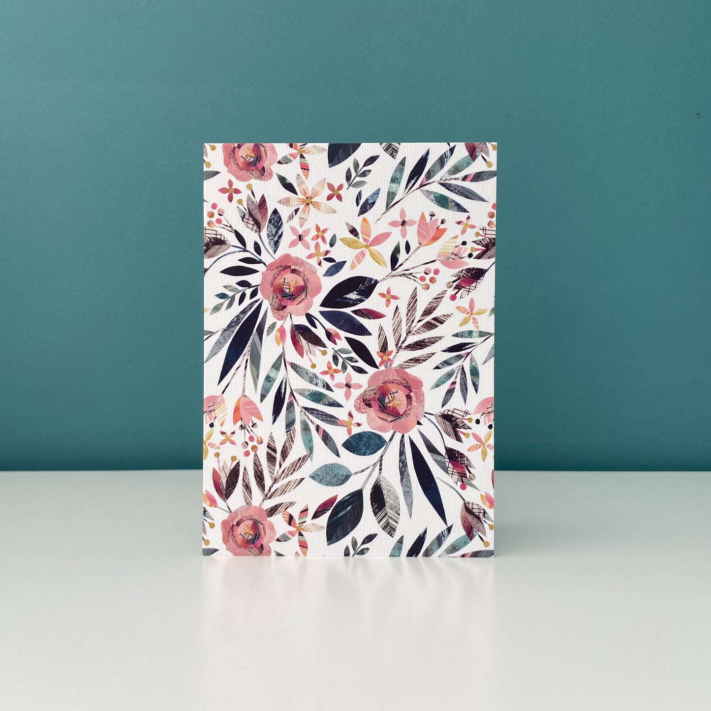 This card features a Summer Floral pattern in pink and green and is reminiscent of a traditional English Cottage garden.  It has been printed onto lightly textured card and is stood on a white shelf against a blue green wall.