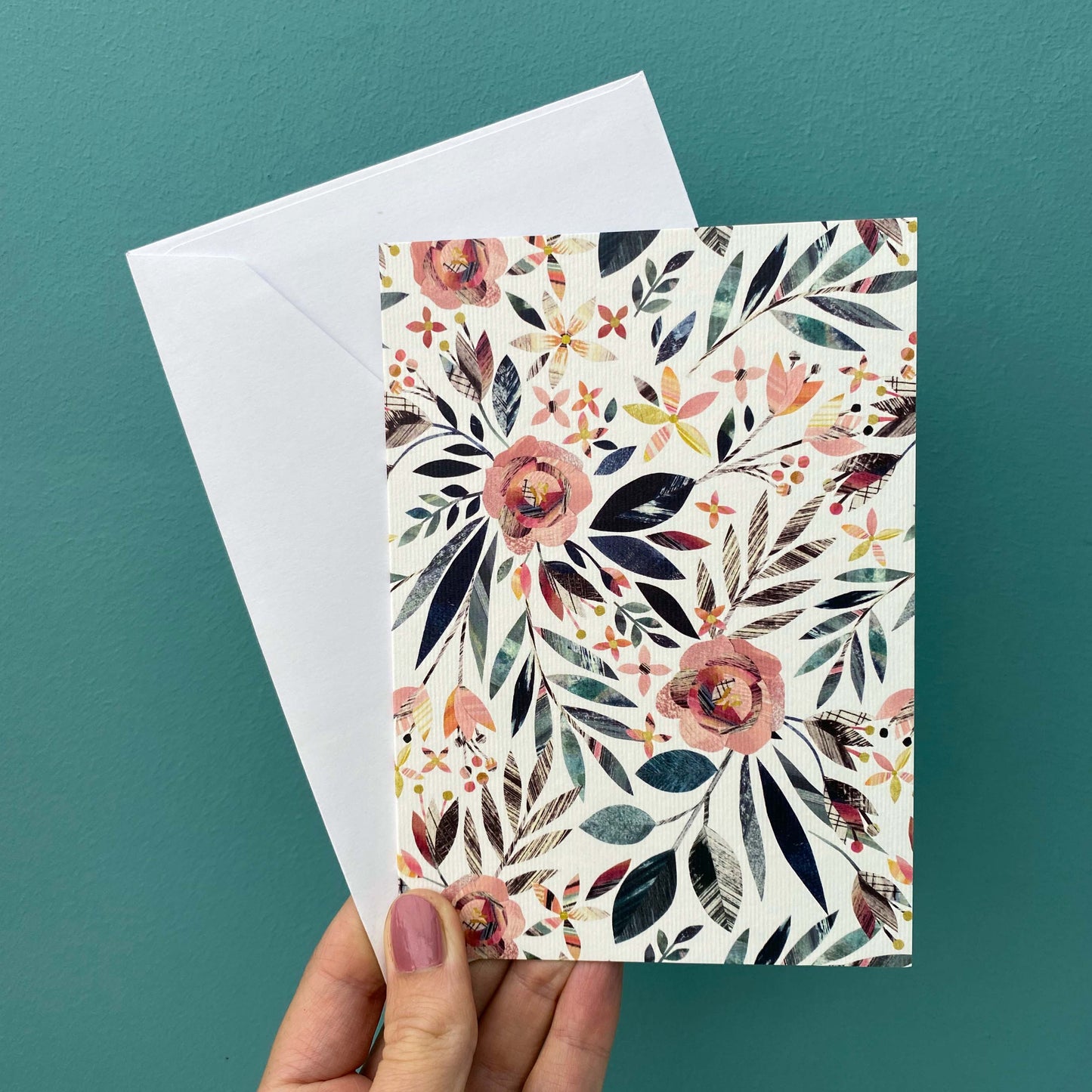 A card featuring an all over pattern of textured collaged dusky pink Roses and Green foliage is being held with a white envelope against a blue green background.