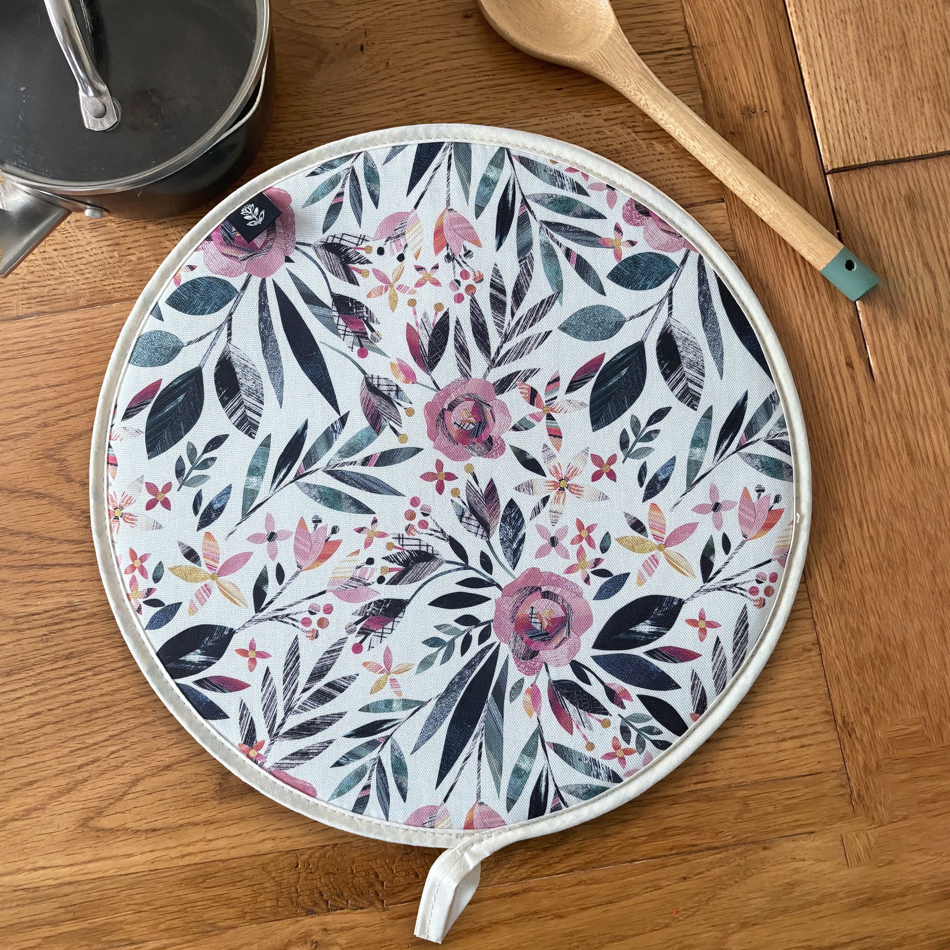 Summer Floral Aga Cover featuring Pink roses and green leaves has been placed on a wooden table with a saucepan and a wooden spoon.