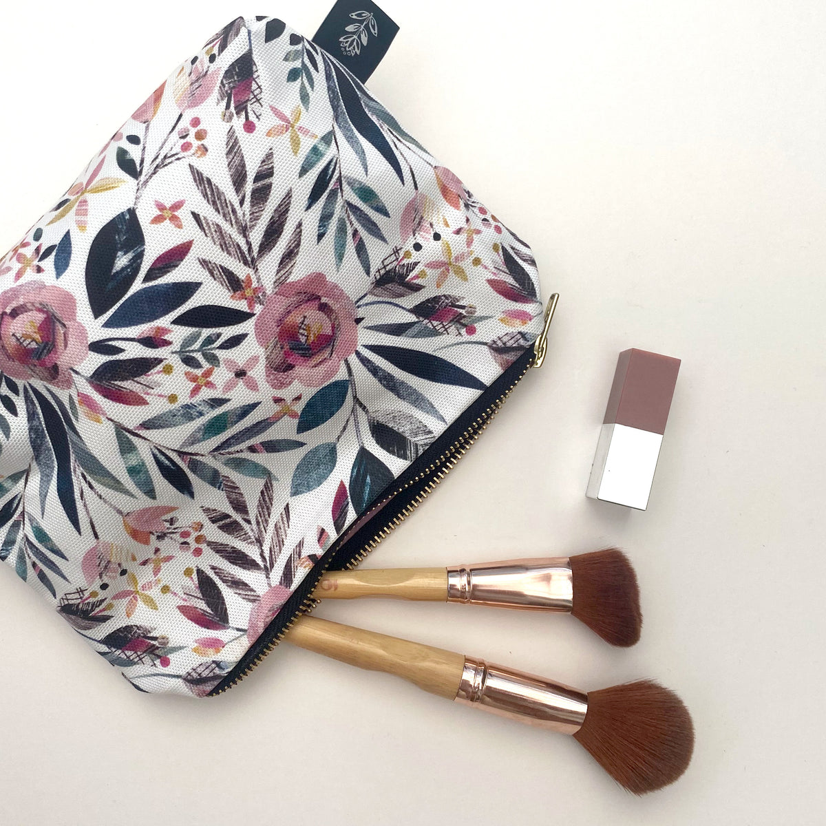 A floral makeup pouch with a gold zipper, shown alongside two makeup brushes and a small lipstick tube on a clean white background.
