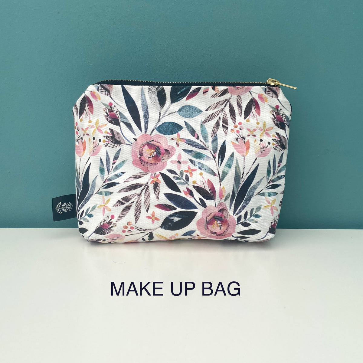 A rectangular makeup bag with a white background and a colorful floral print featuring pink, teal, and dark blue flowers and leaves. The bag has a gold zipper and a small black fabric tag on the left side.