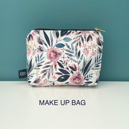A rectangular makeup bag with a white background and a colorful floral print featuring pink, teal, and dark blue flowers and leaves. The bag has a gold zipper and a small black fabric tag on the left side.