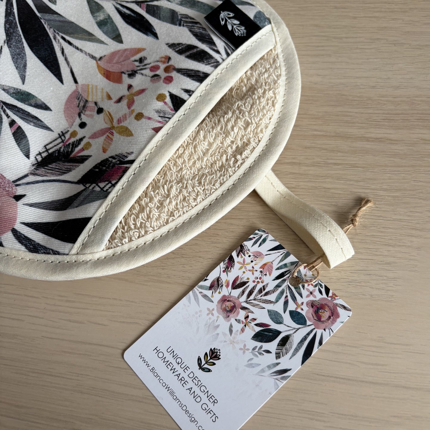 Summer Floral pot holder has been placed on a wooden table and has a swing tag tied around its hanging hook with twine.  The brand label can also been seen on the opening for the hand.