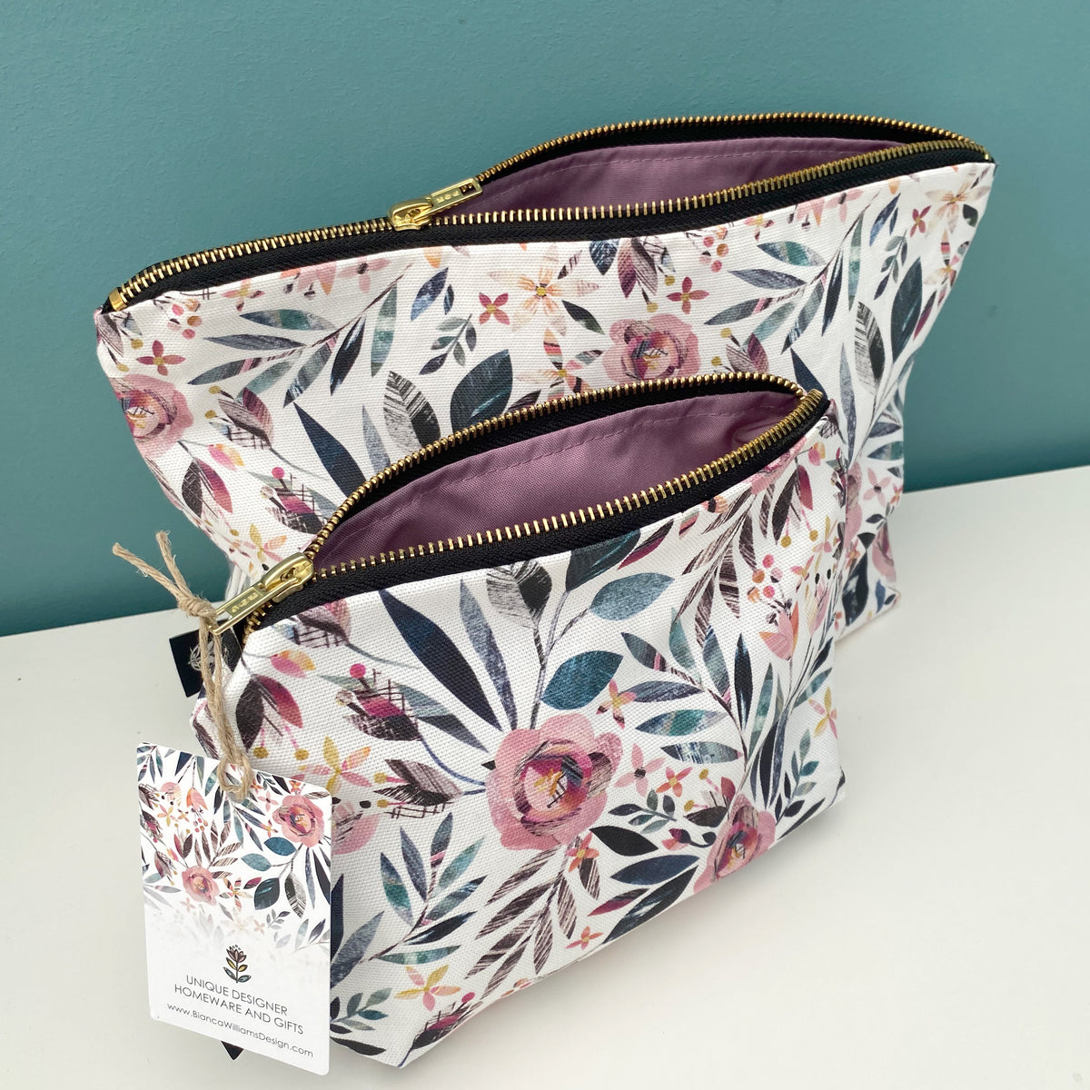 Two floral zippered pouches with gold zippers and a purple interior lining, displayed on a white surface against a teal background. A paper tag is attached to the front pouch.