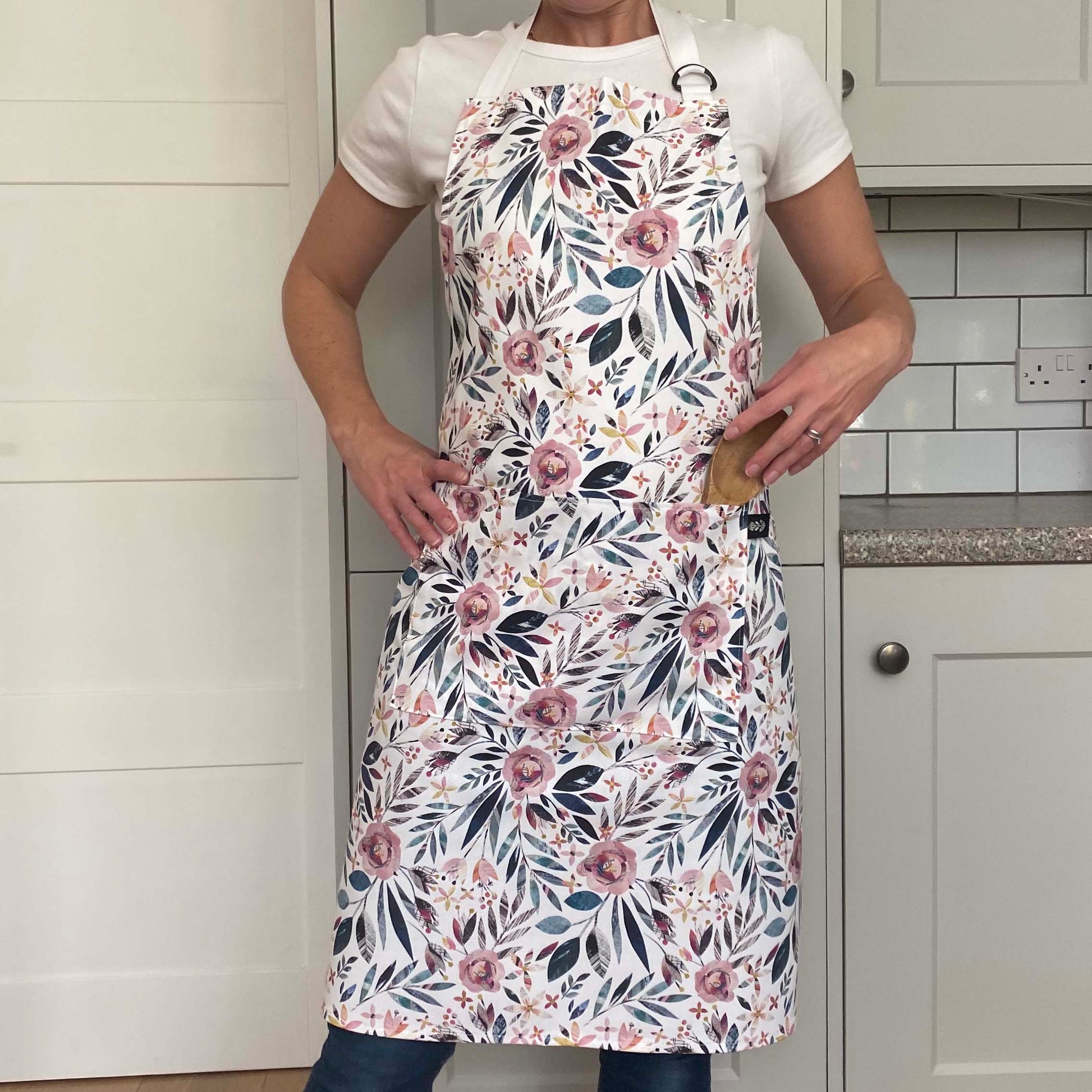 The model wears a Summer Floral Apron featuring pink roses with green foliage.  The apron has two front pockets and an adjustable white neck strap.
