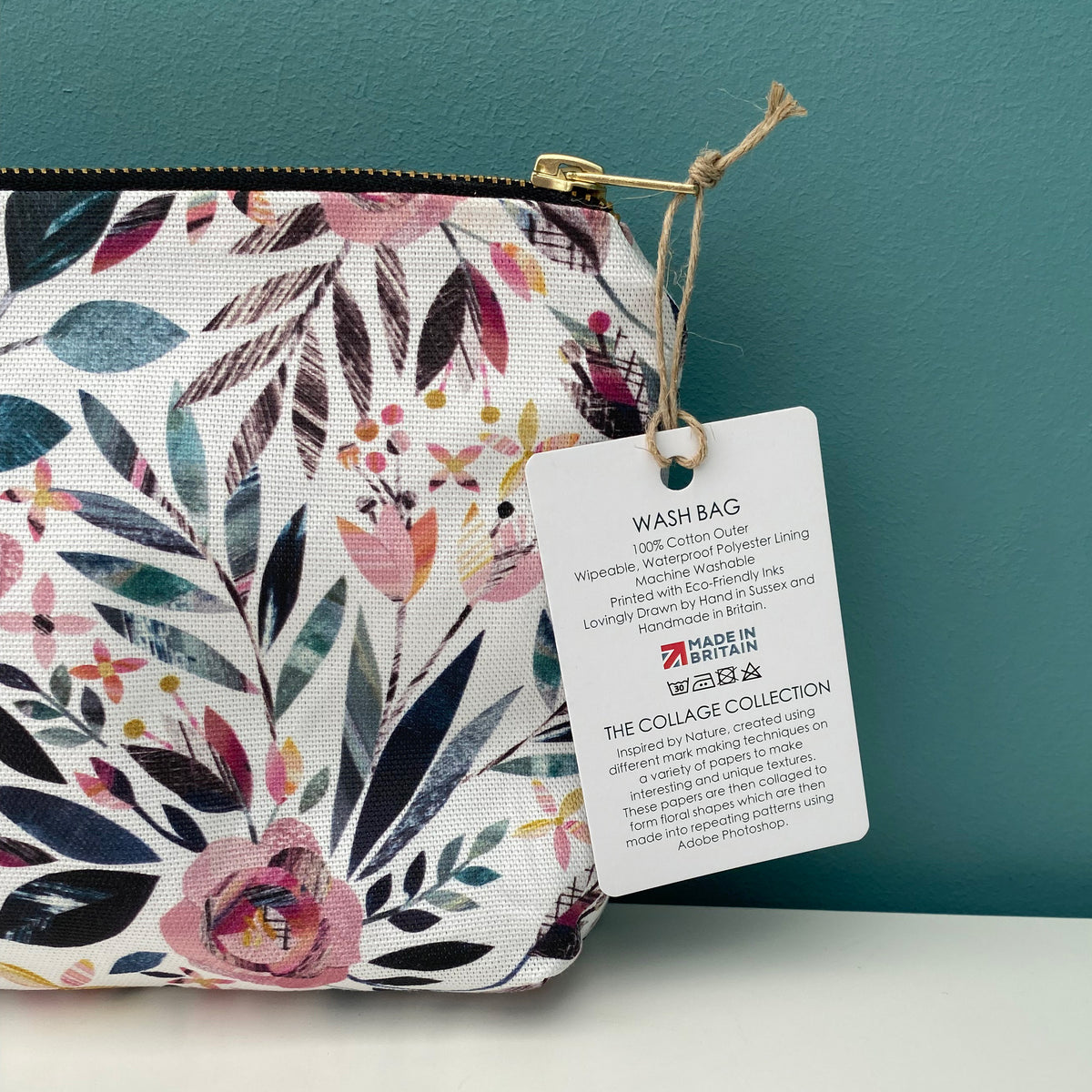 A close-up of a fabric wash bag with a colorful floral and leaf print on a white background, featuring a gold zipper and a hanging product tag that reads 'WASH BAG' with 'The Collage Collection' branding.