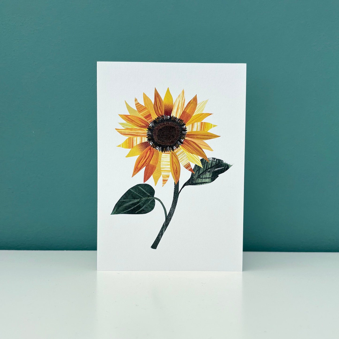 White greeting Card with a sunflower design stood on a white surface against a teal background.
