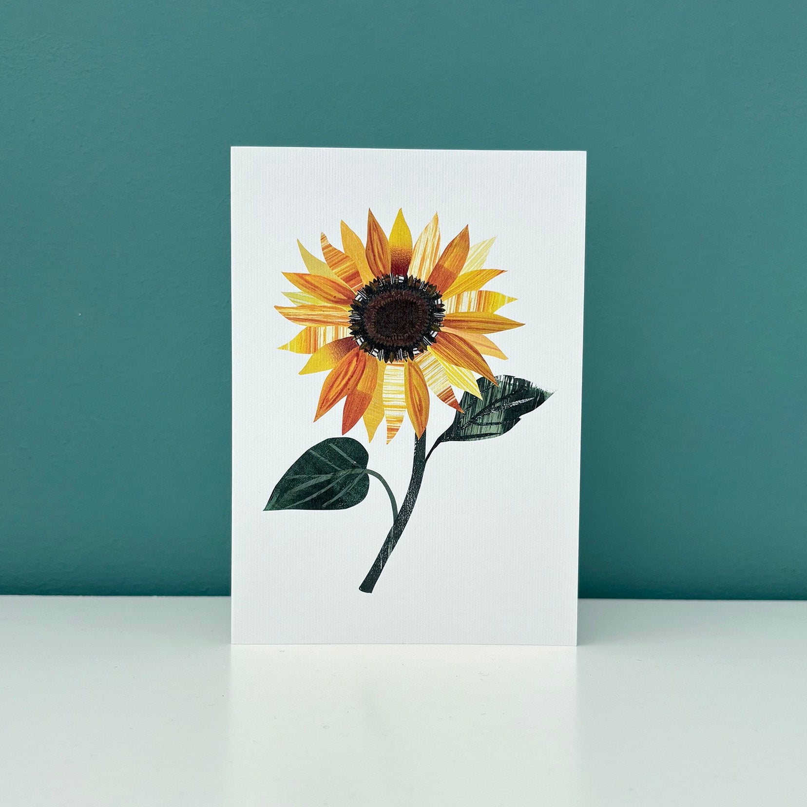 White greeting Card with a sunflower design stood on a white surface against a teal background.