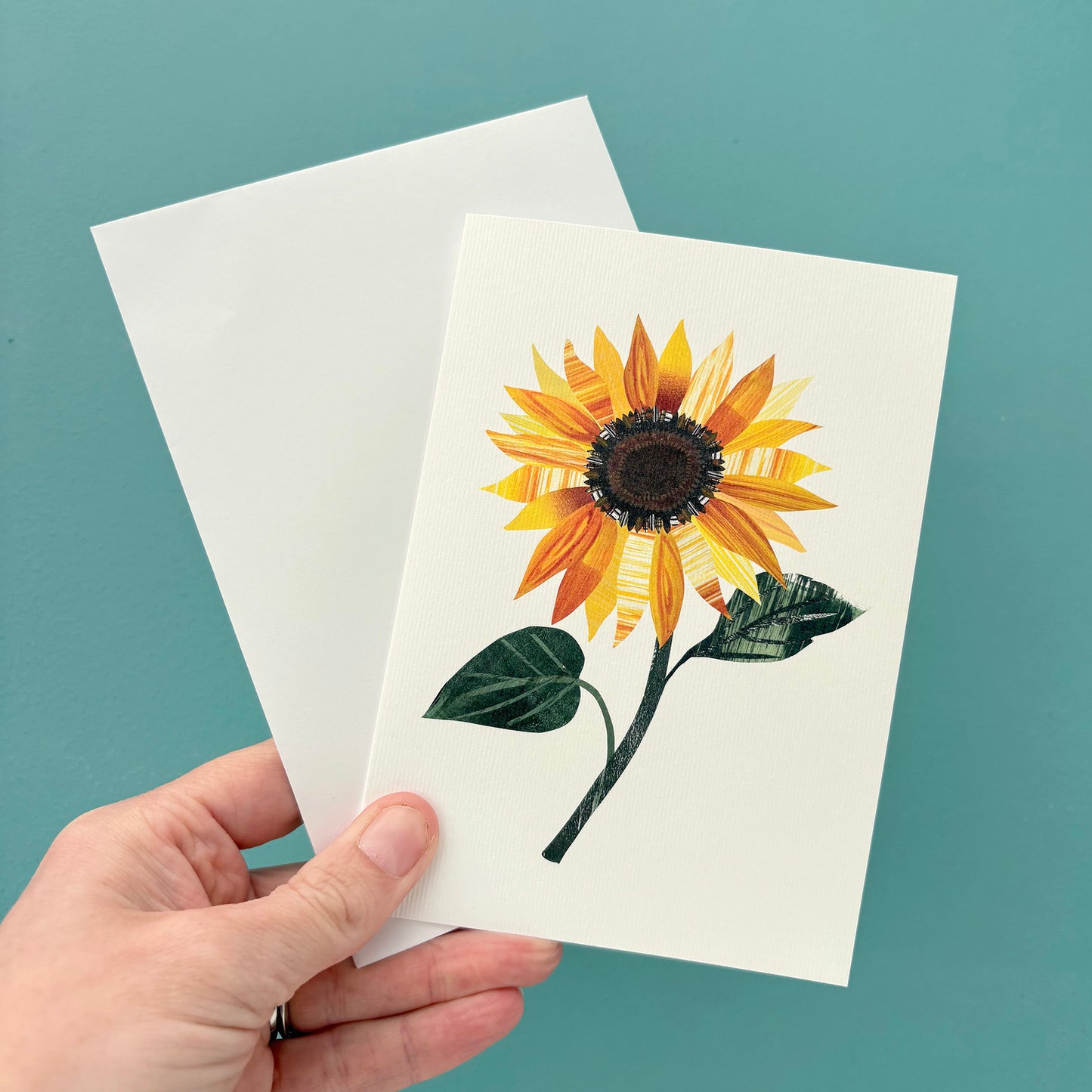 Hand holding a card with a sunflower design and a white envelope against a teal background.