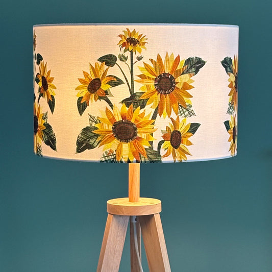 A large Sunflower lampshade has been placed on a wooden lamp base and has been switched on, it is against a blue green background.