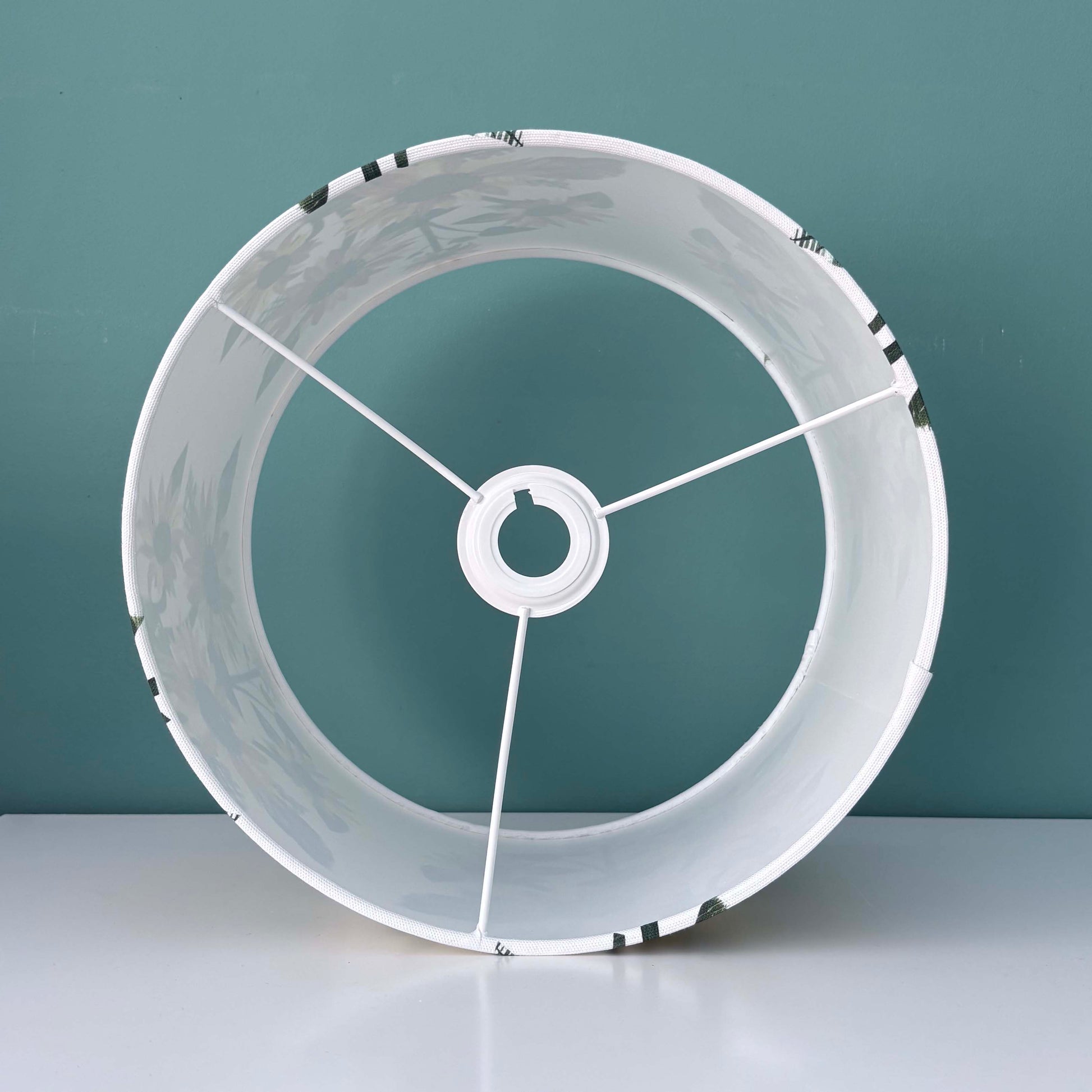 A sunflower lampshade has been placed on its side on a white shelf, so that you can see the lining of the lampshade.  It is against a blue green wall.