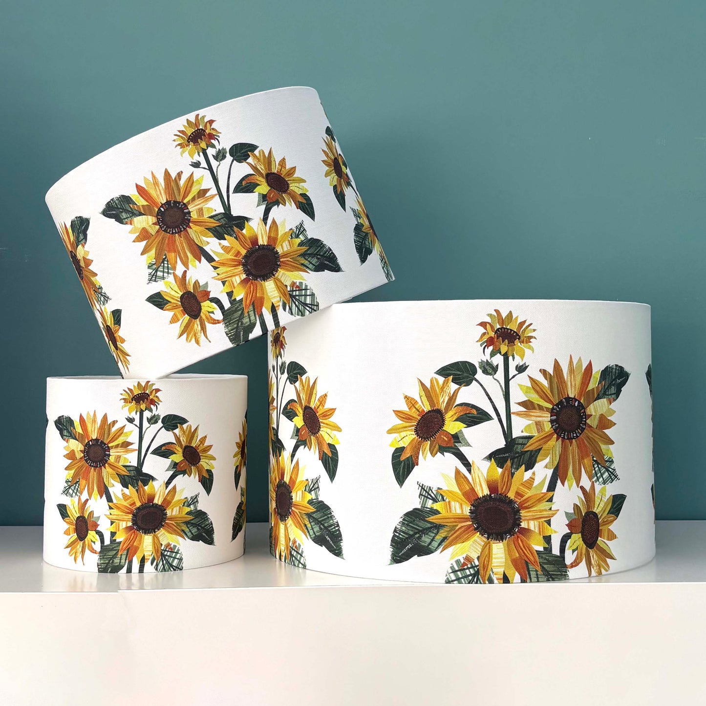 Three Sunflower Lampshades in three different sizes have been placed on a white shelf against a blue green background.