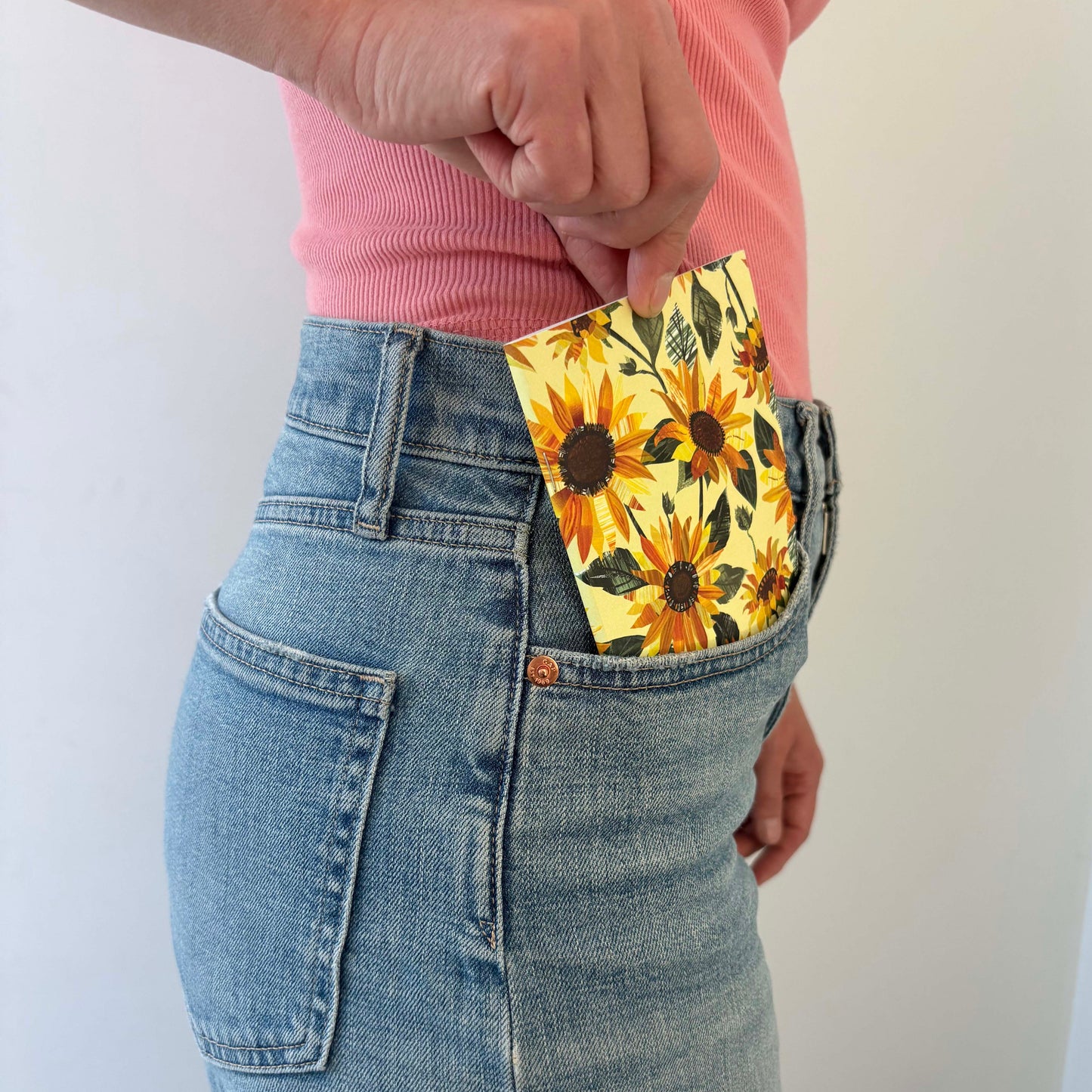 A Sunflower Notebook can be seen being placed in the front pocked of a ladies blue jeans.