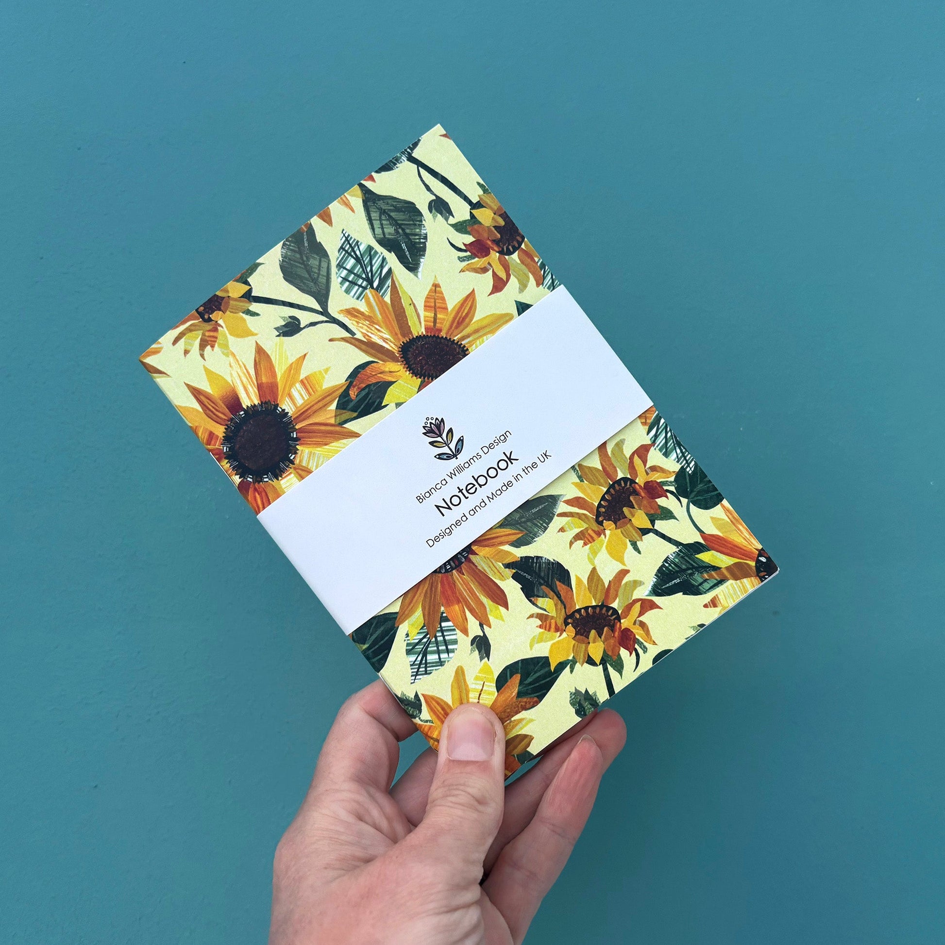 Small yellow notebook with all over Sunflower pattern packaged in a white branded belly band and held in front of a blue green back ground.