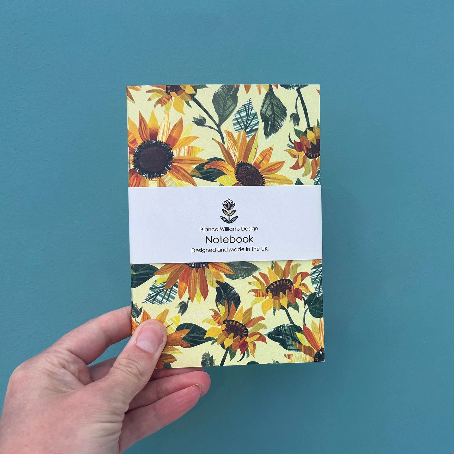 Small yellow notebook with all over Sunflowers pattern packaged in a white branded belly band and held in front of a blue green back ground.