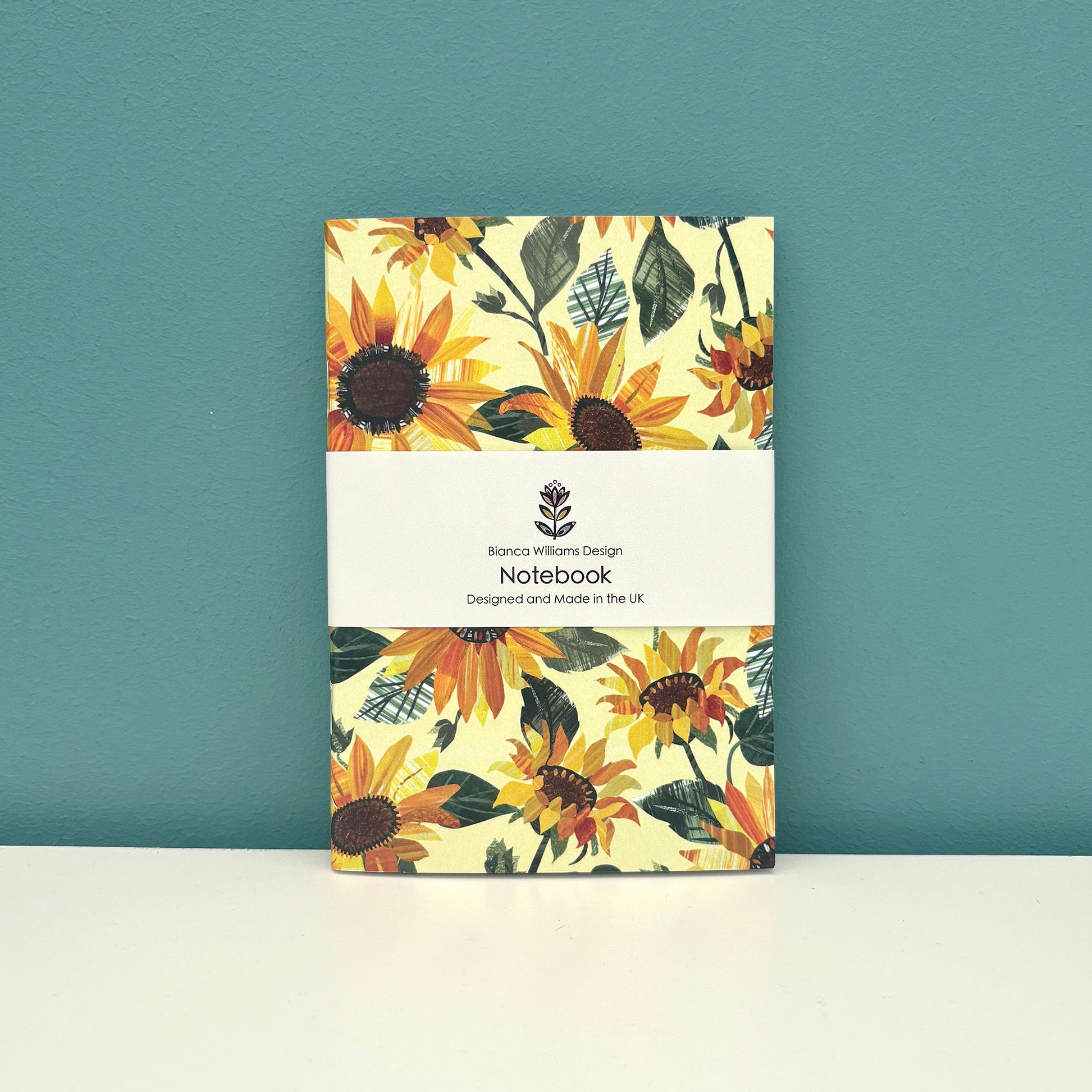 A small yellow Notebook covered in an all over Sunflower pattern has been packaged in a branded belly band and has been placed on a white shelf with a blue green wall behind it.