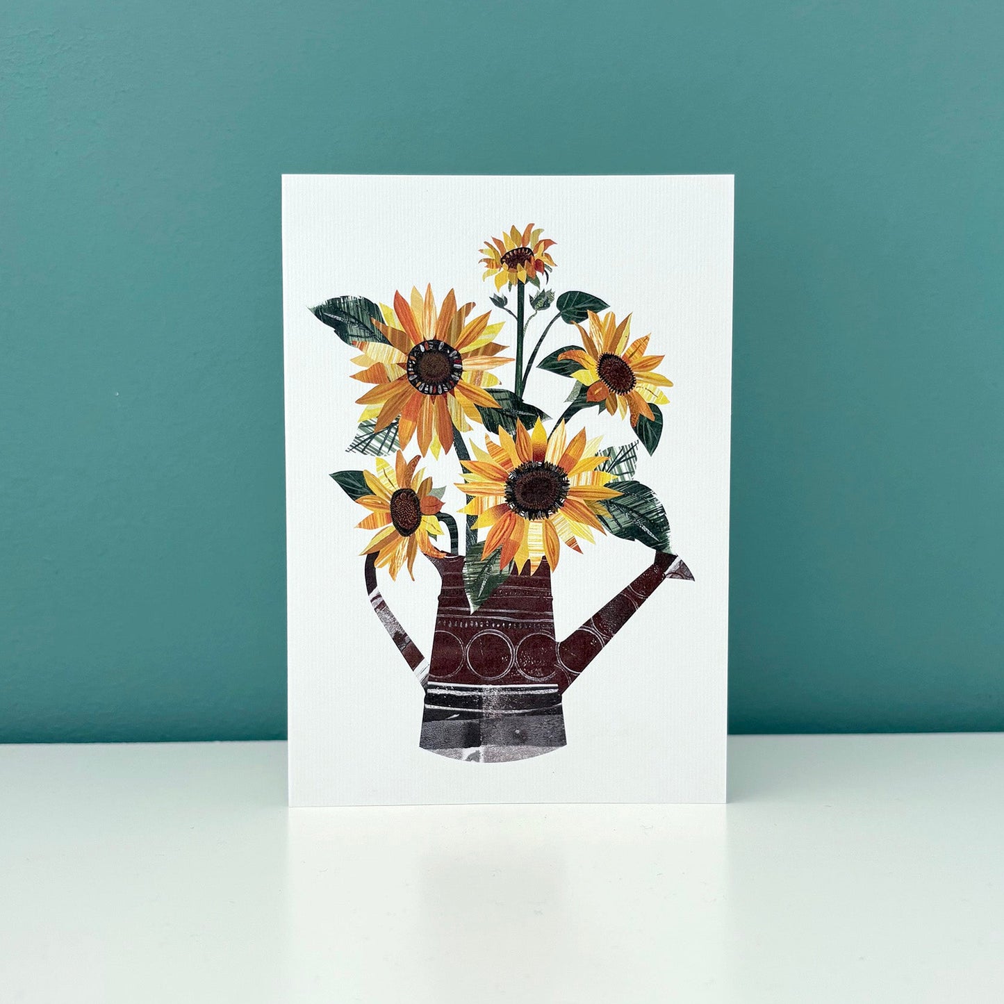 A white card with an image of yellow and Orange sunflowers with green leaves in a watering can has been stood on a white shelf against a blue green wall.