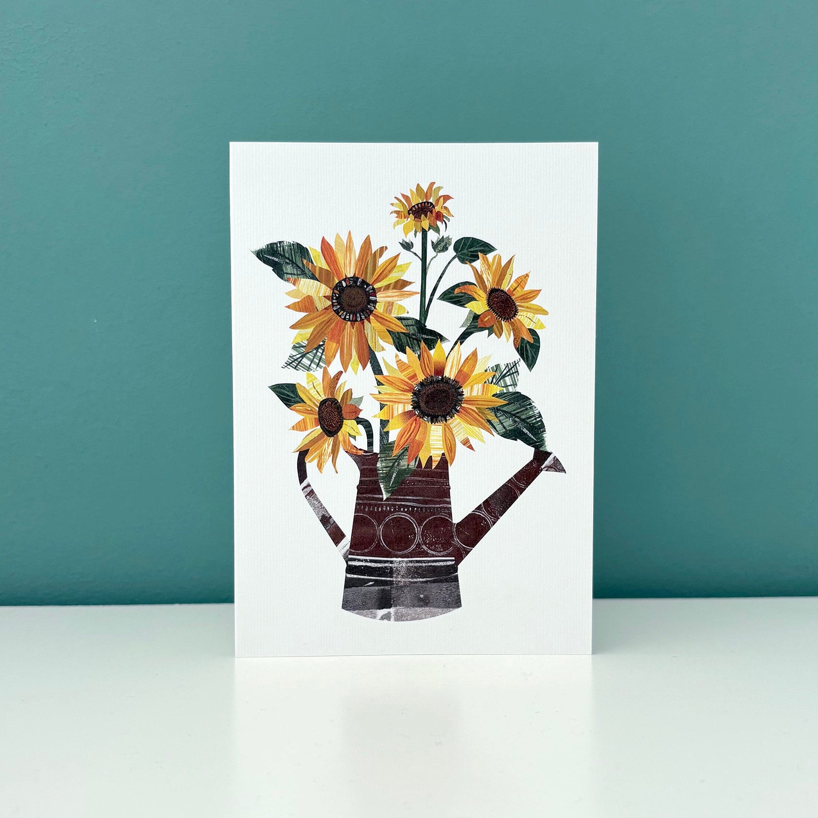 A white card with an image of yellow and Orange sunflowers with green leaves in a watering can has been stood on a white shelf against a blue green wall.