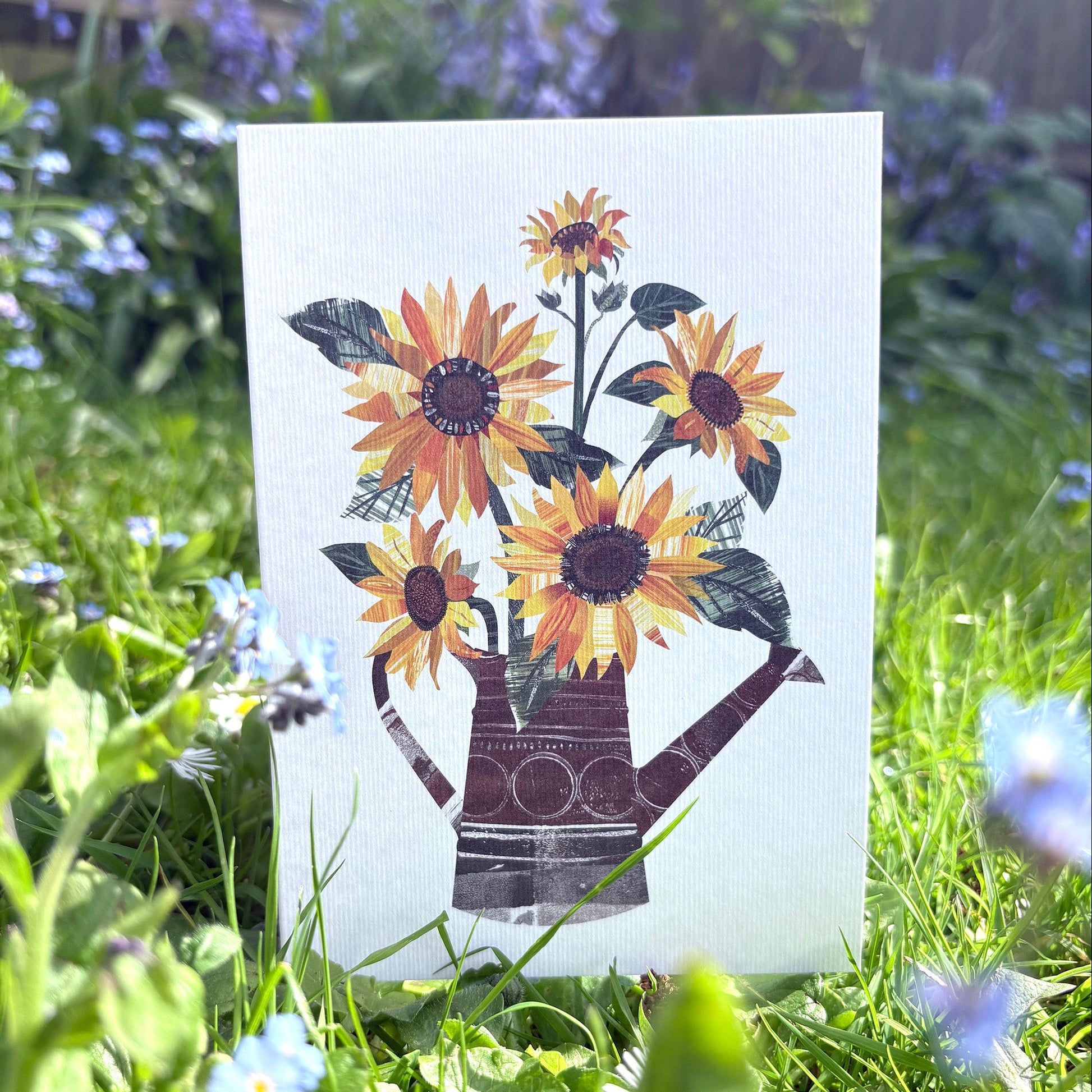 A white card with yellow and orange sunflowers with green leaves in a watering can has been stood up on some grass outside.