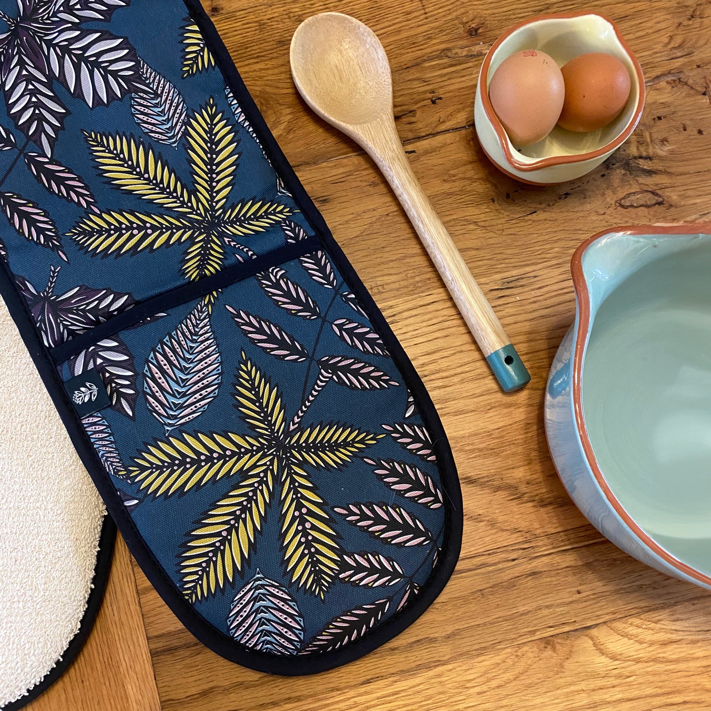 Teal Forest Floor Double Oven Gloves have been placed at an angle and laid onto an oak table.  A wooden spoon and two bowls have also been laid onto the table.  You can see the Brand Label sewn into the outside seam of the oven gloves.  You can also see the Ecru coloured cotton towelling on the reverse of the Oven Gloves.