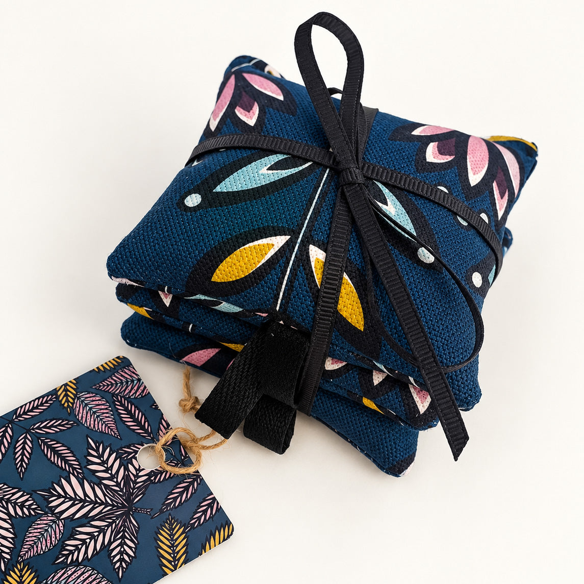 A pack of three lavender bags have been tied with black ribbon, they have a dark teal background and feature a stylised flower motif in Black, white, aqua, yellow and ducky pink they have been placed on a plain white background.