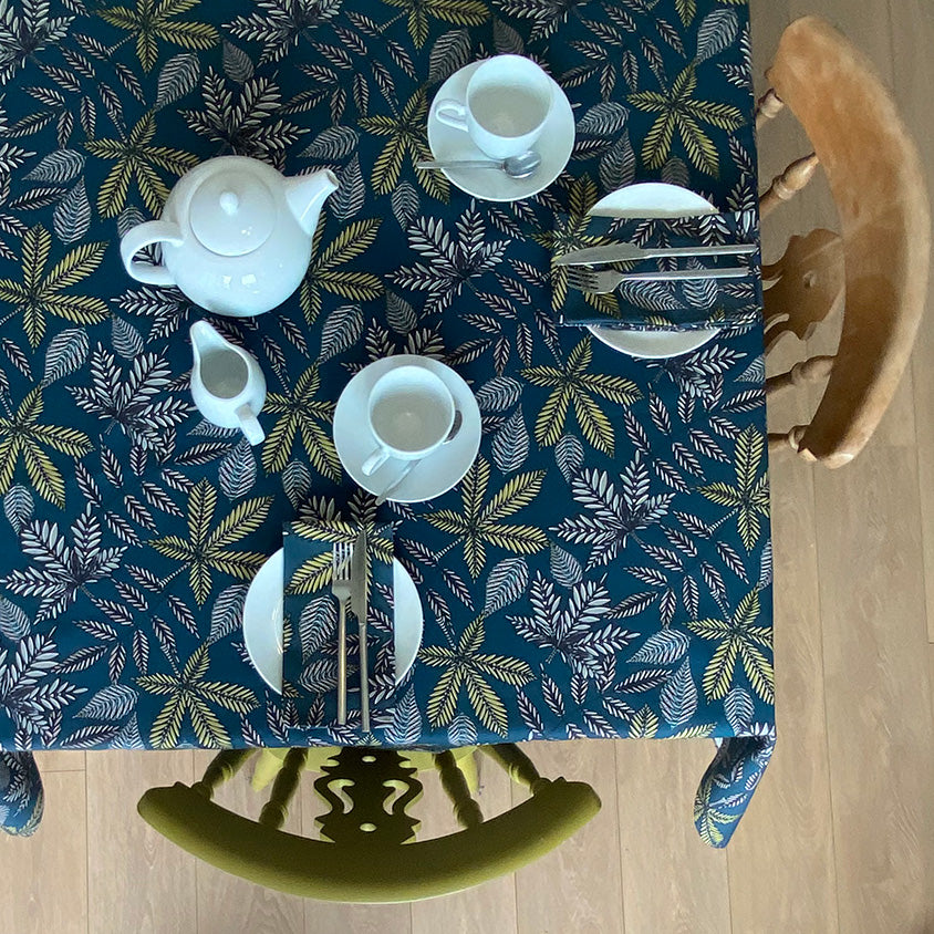 Teal Forest Floor Table Cloth from a Birds Eye view.  The Teal Tablecloth features  different shapes leaves in Black, Blue, yellow and Grey.  The table is set for two people with small white plates a tea pot and cups. 