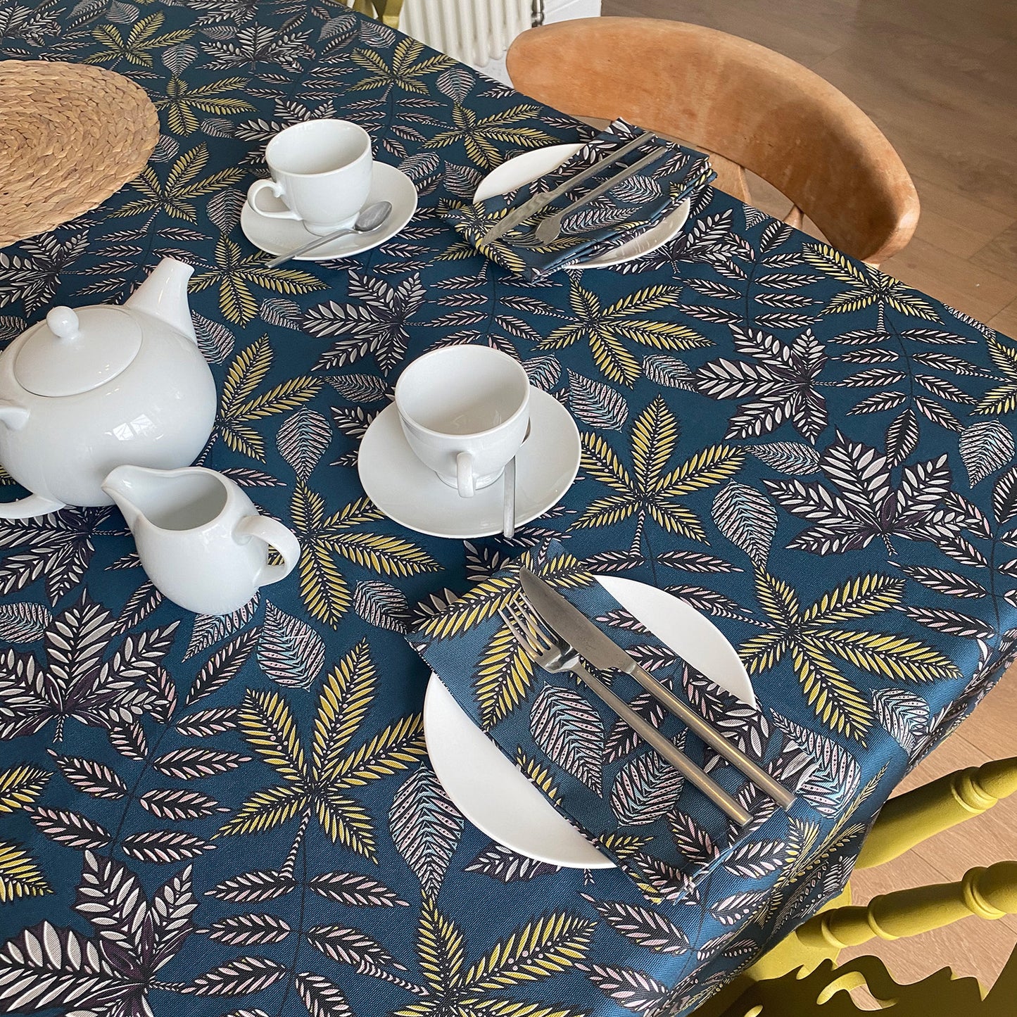 Teal Forest Floor Table Cloth from an angle. The Teal Tablecloth features different shapes leaves in Black, Blue, yellow and Grey. The table is set for two people with small white plates a tea pot and cups.