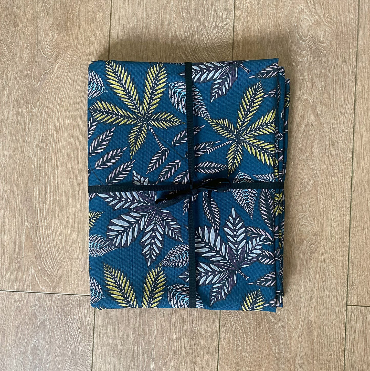 The Teal Forest Floor Table cloth has been folded and is packaged tied up in a bow with black cotton ribbon.