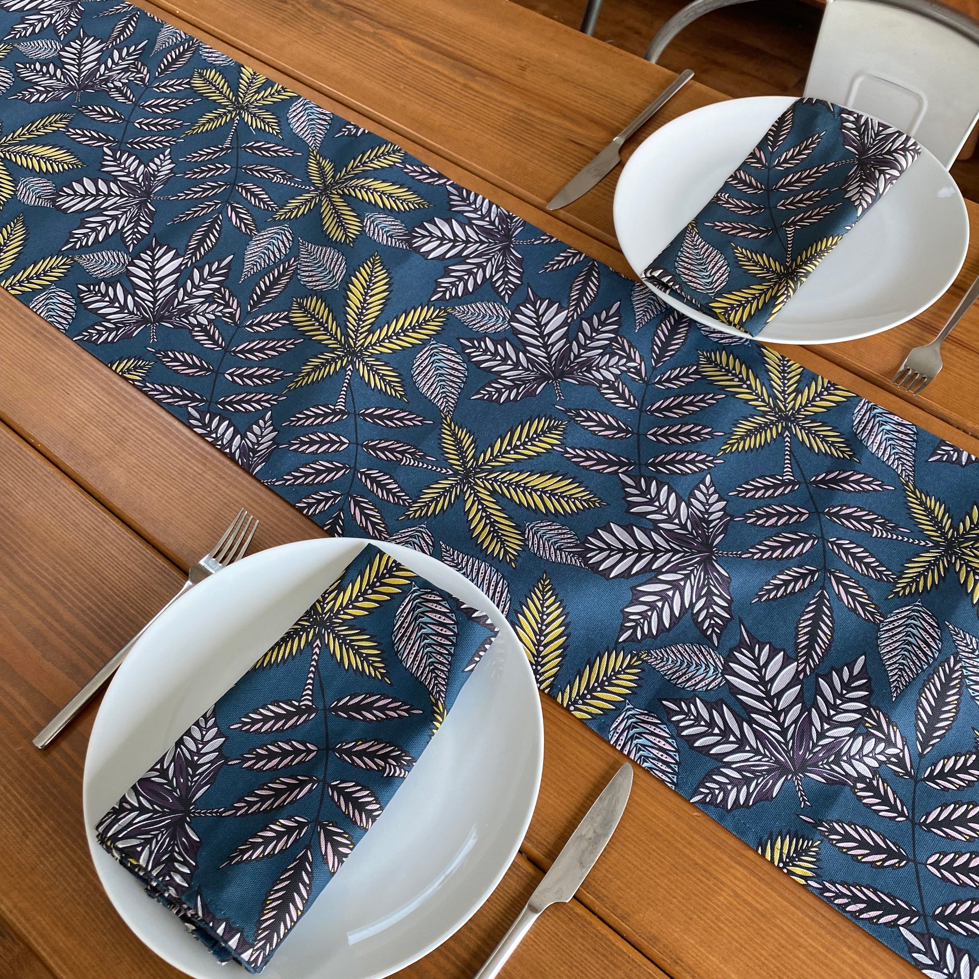 A teal Forest Floor table runner has been placed down the middle of a wooden table.  The table runner has different shaped leaf motifs on it in Black, white, yellow, blue and dusky pink printed onto a dark Teal green.  The table has been set with two plates and two matching napkins.