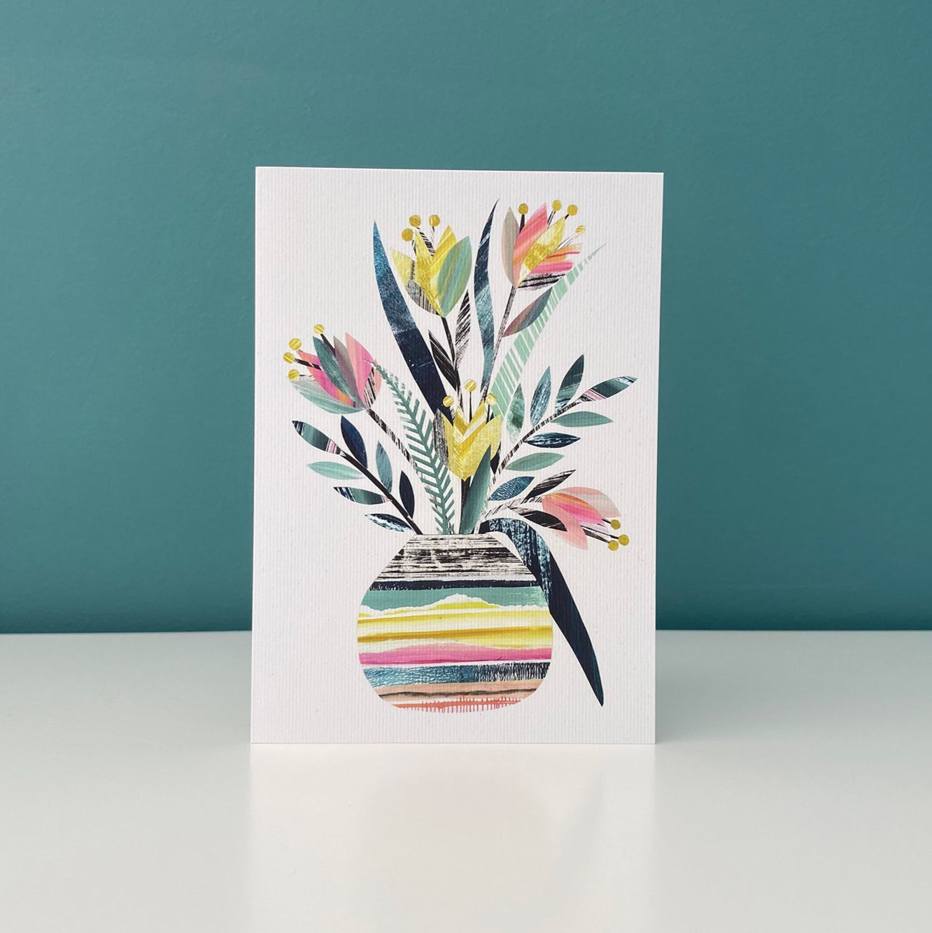 A tulip Vase card featuring colourful pretty Pink, green and yellow tulip flowers with green leaves have been stood on a plain white shelf with a blue green back ground.