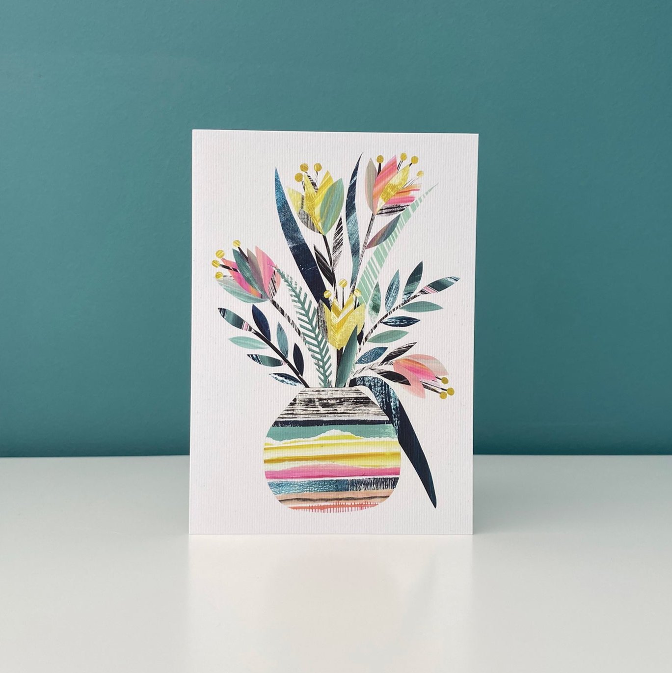 A greeting card featuring a colourful vase of stylised Tulips in yellows, pinks and green on a white card has been stood on a white shelf with a blue green background.