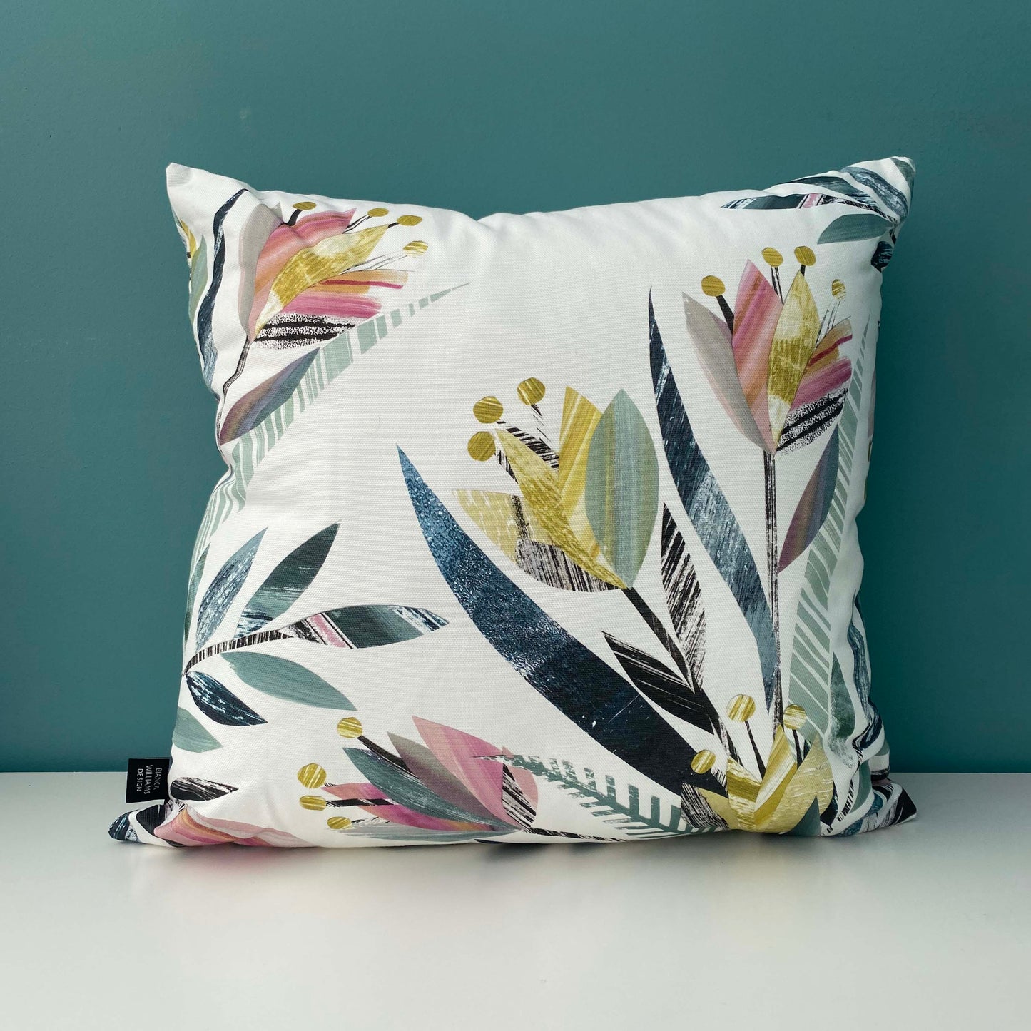 A Square Tulip Cushion featuring a textured tulip pattern in Pink, yellows, greens and grey on a white background has been placed on a white shelf against a blue/green wall.
