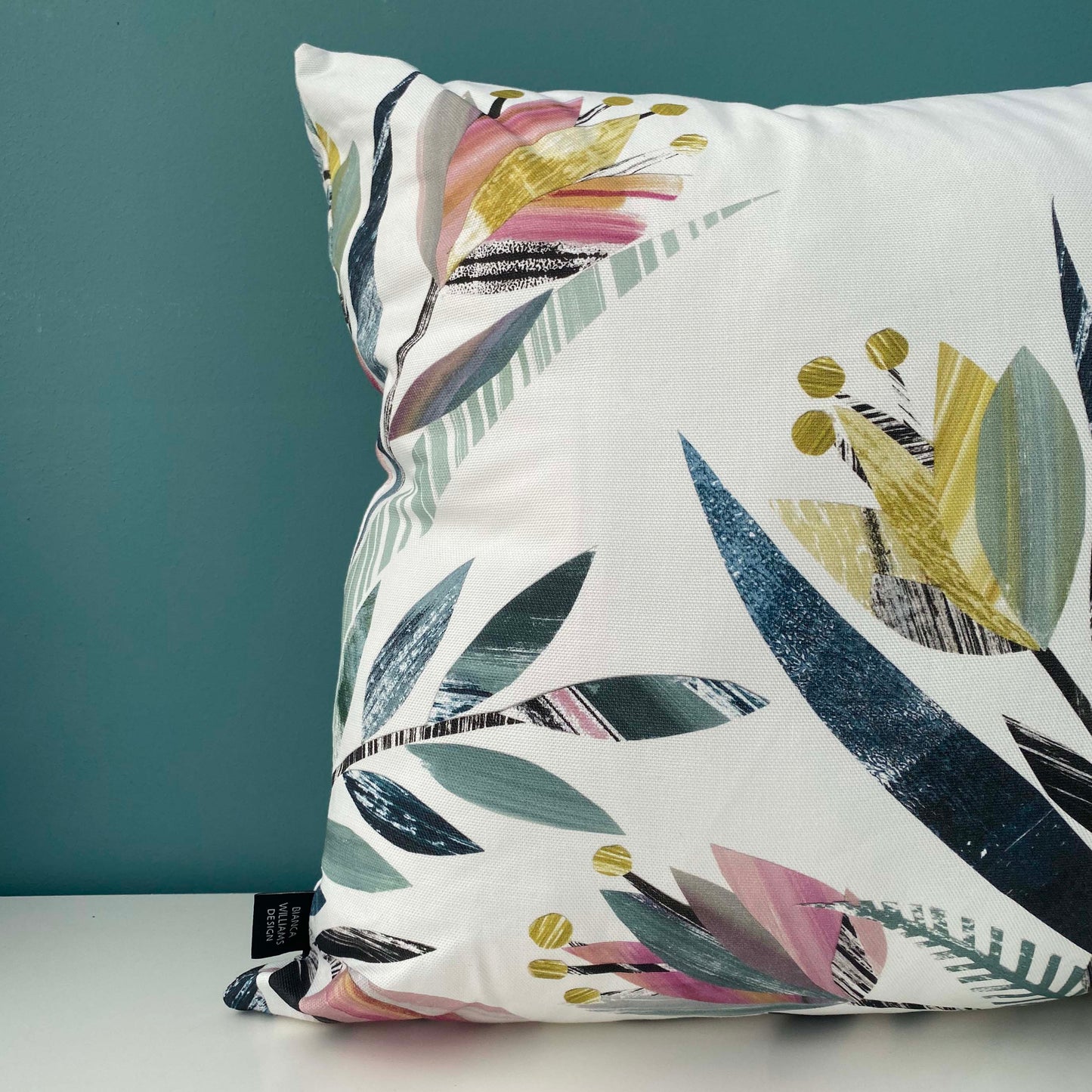 A close up of a Square Tulip Cushion featuring a textured tulip pattern in Pink, yellows, greens and grey on a white background has been placed on a white shelf against a blue/green wall.