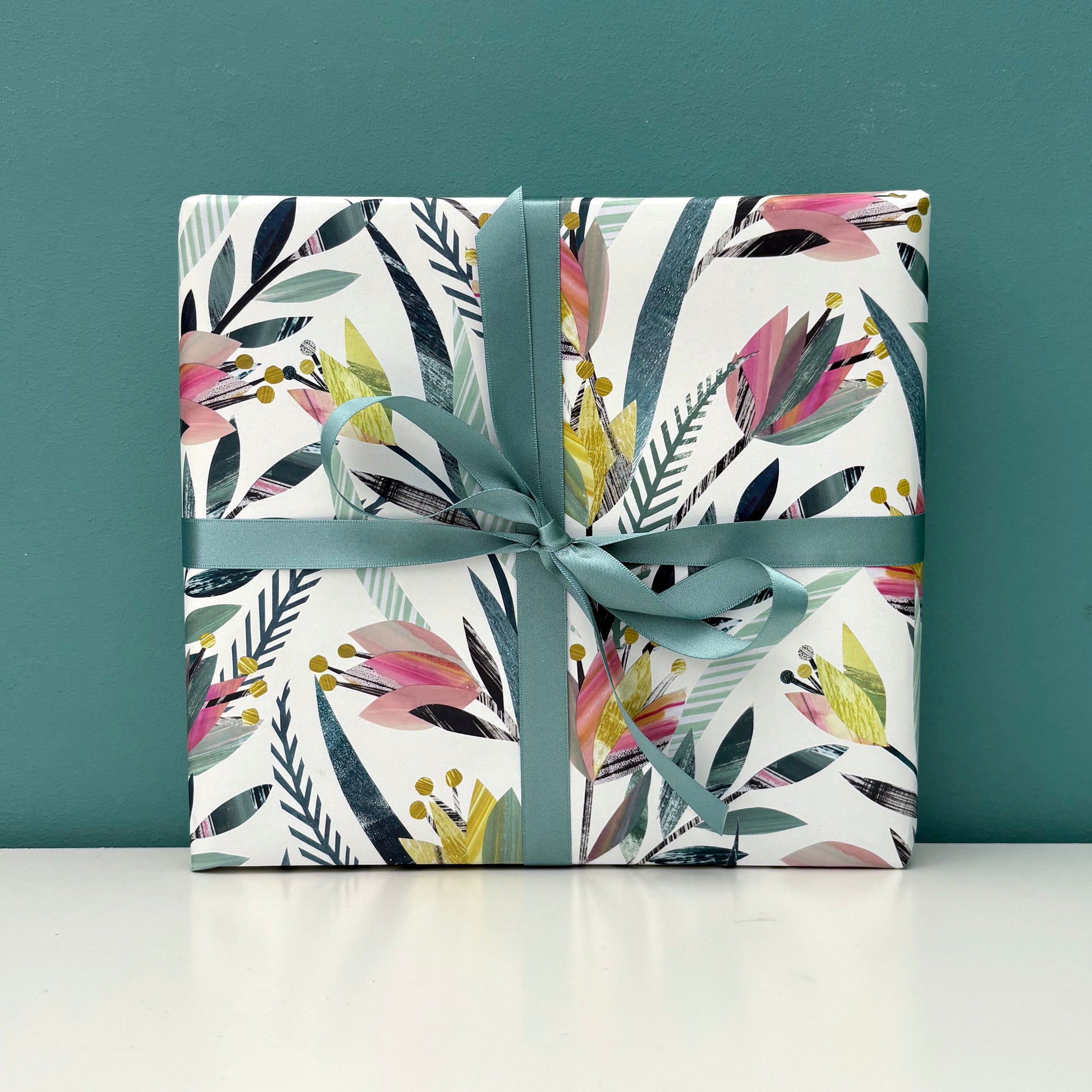 A gift wrapped in tulip floral wrapping paper tied with coordinating green ribbon has been placed on a white shelf against a blue green wall.