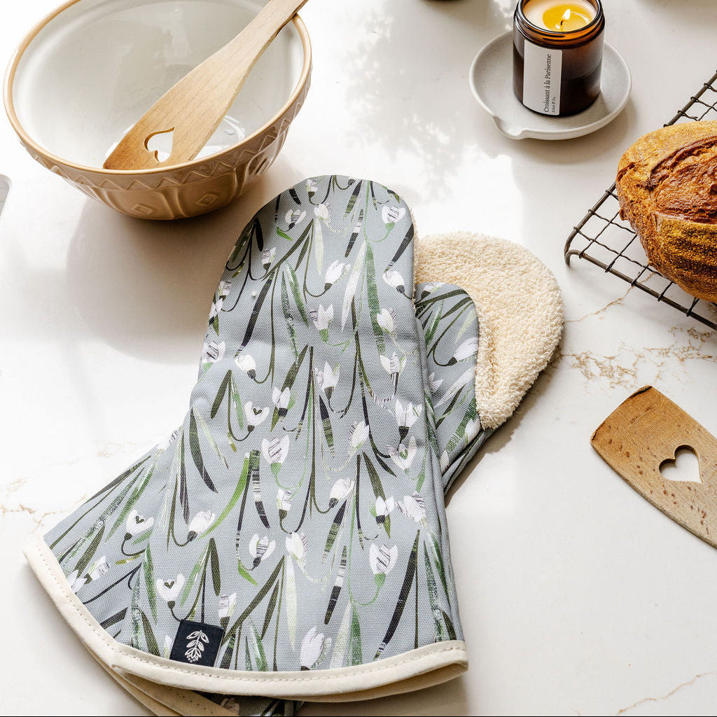 A Snowdrop patterned kitchen oven mitt with a green grey background, white Snowdrops, and green stems, featuring a beige cotton towelling lining, styled on a white marble countertop with a ceramic bowl, wooden spoon, candle, and bread.