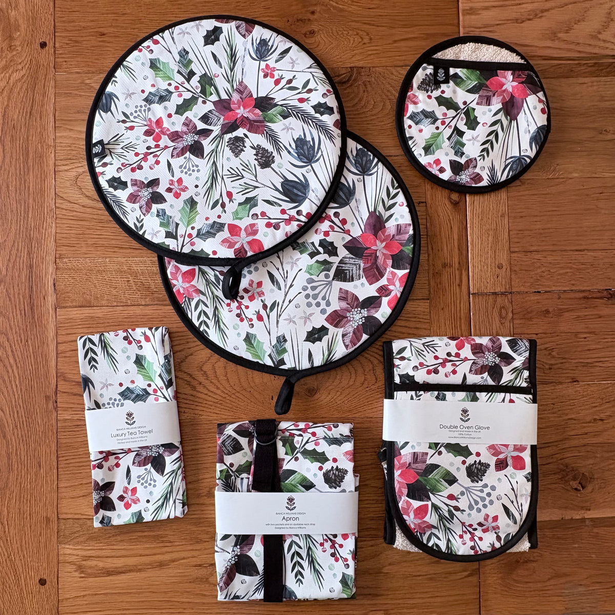 A Winter Floral kitchen textile set arranged on a wooden table and includes two round Aga Covers, a pair of oven mitts, a tea towel, an apron and a pot grab all in the same matching floral print. The fabric has a white base with red Poinsettia Flowers, green Holly and red berries.